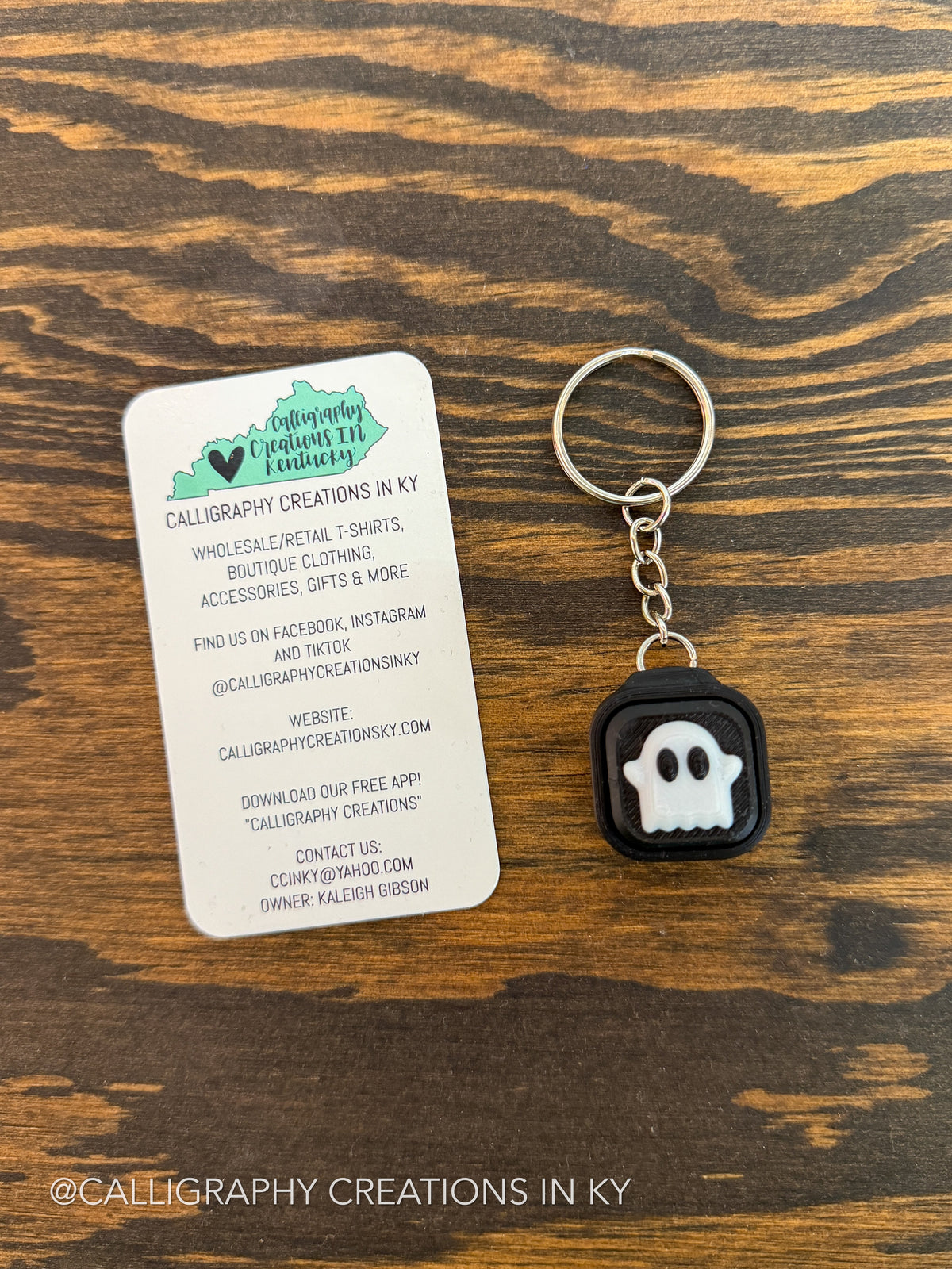 A small black square keychain with a white ghost face and silver key ring, placed beside a white branded card on a dark wood surface.