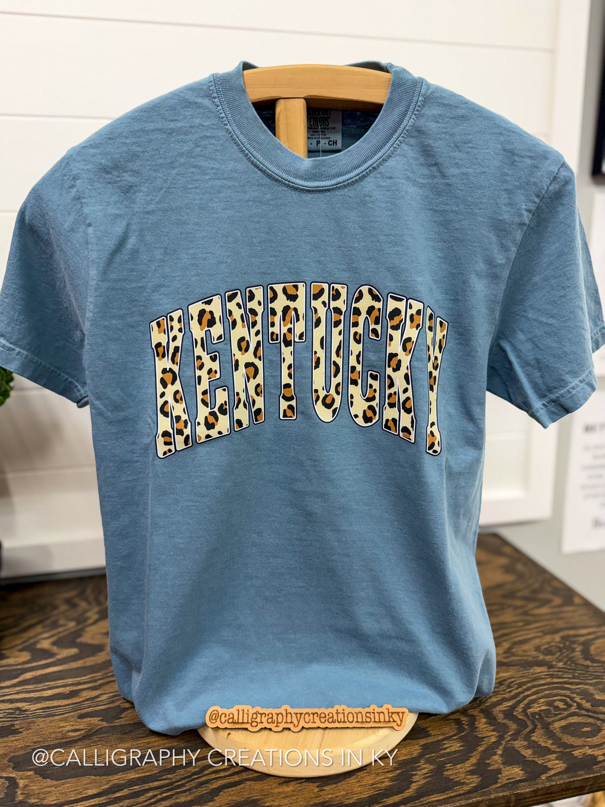A light blue crewneck t-shirt on a wooden hanger, featuring the word 'KENTUCKY' printed across the chest in an arched leopard-print pattern.
