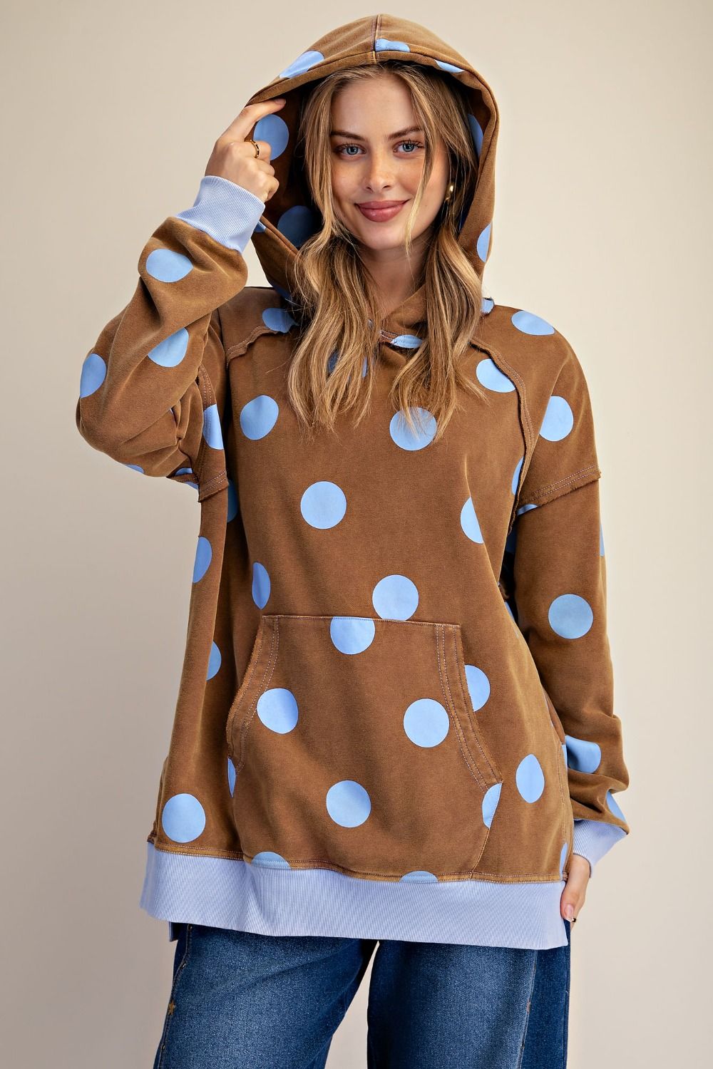 A woman wearing a brown hooded sweatshirt with large light blue polka dots and light blue ribbed trim, paired with blue jeans, against a plain beige studio background.