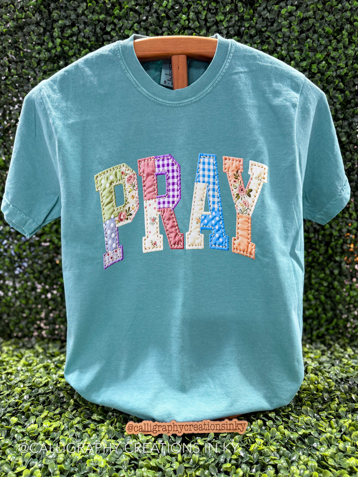 A turquoise short-sleeve t-shirt with the word 'PRAY' spelled out in colorful patchwork letters, hanging on a wooden hanger against a green hedge background.