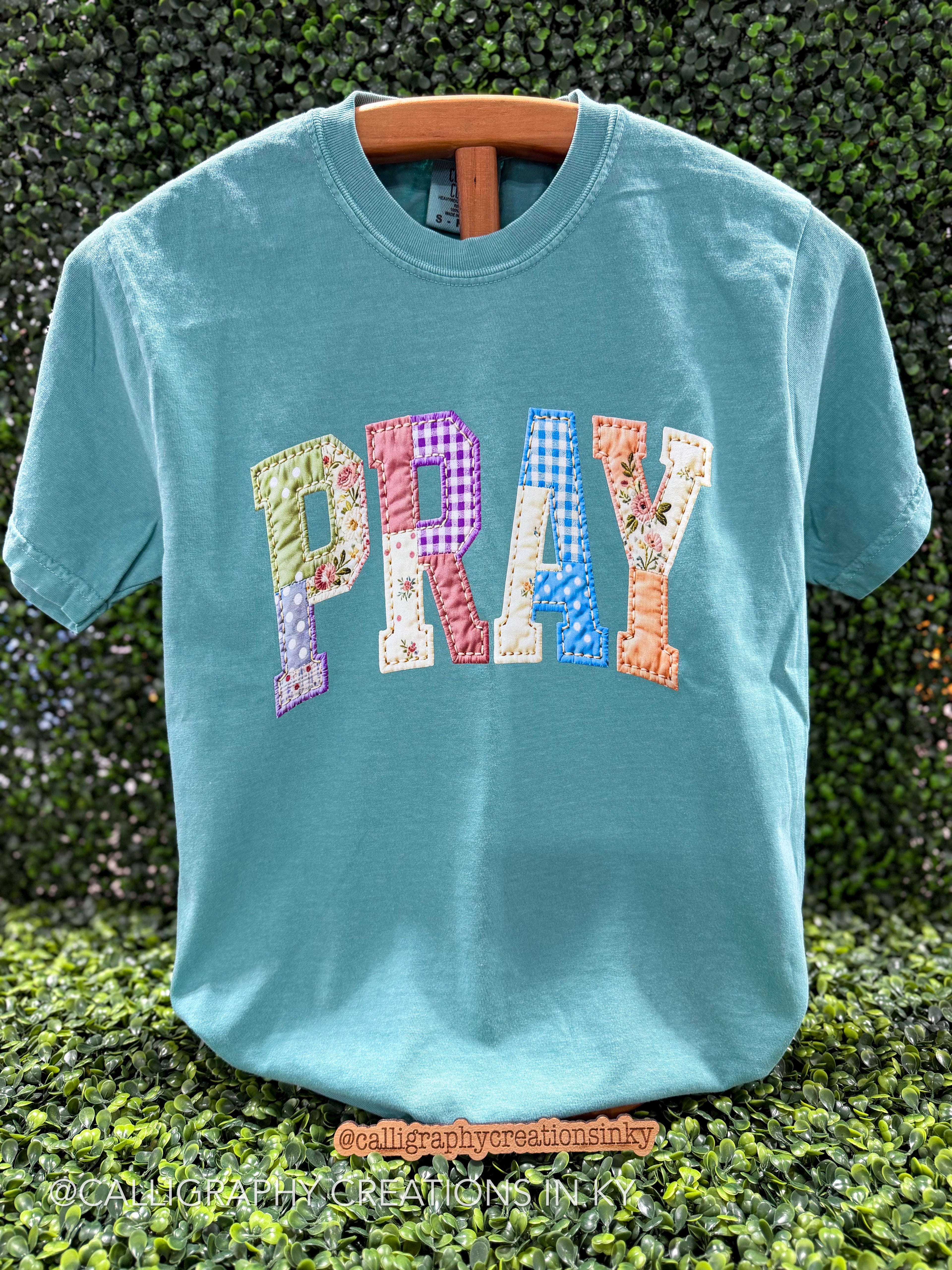 A turquoise short-sleeve t-shirt with the word 'PRAY' spelled out in colorful patchwork letters, hanging on a wooden hanger against a green hedge background.