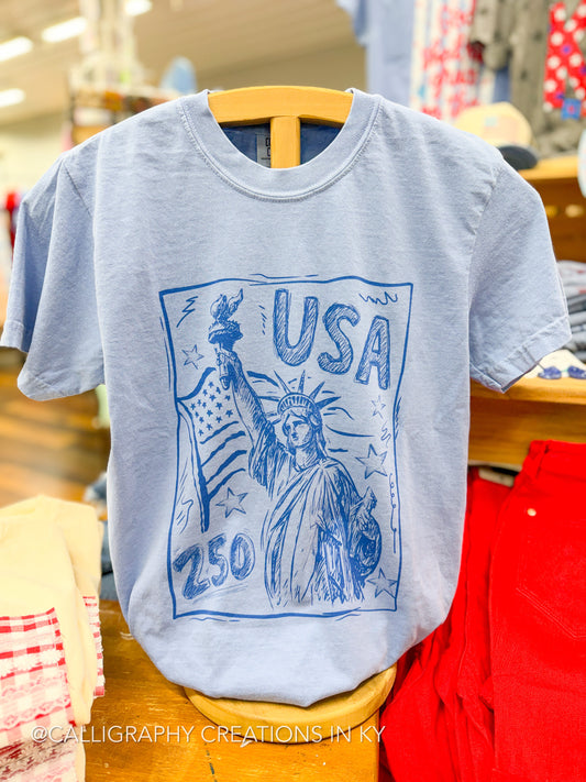 Light blue t-shirt with Statue of Liberty graphic and 'USA 250' text on a hanger.