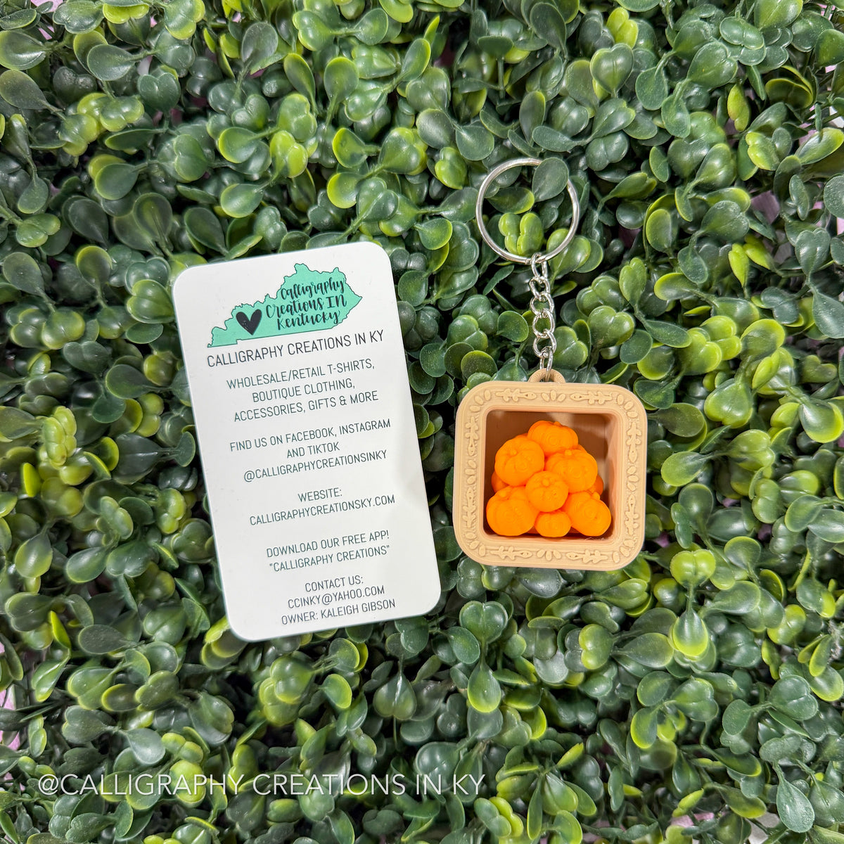 A square wooden calligraphy keychain with a silver metal ring and chain, featuring a light wood tray filled with small orange calligraphy characters, displayed on a background of green leaves next to a white brand card.