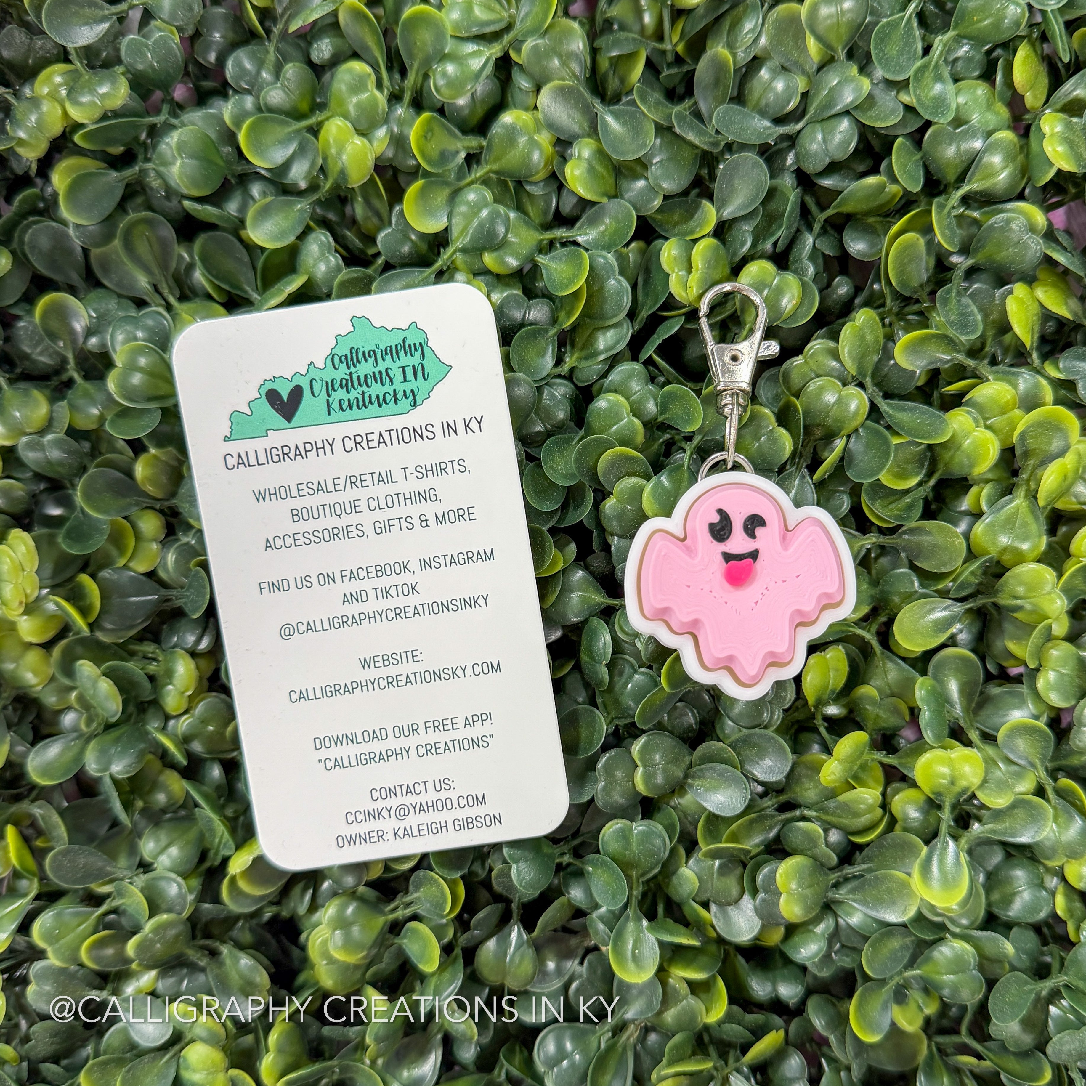 A pink cartoon ghost keychain with a silver clasp is displayed on a white business card, which is placed on a background of green leafy foliage. The card shows the brand 'Calligraphy Creations in KY'.