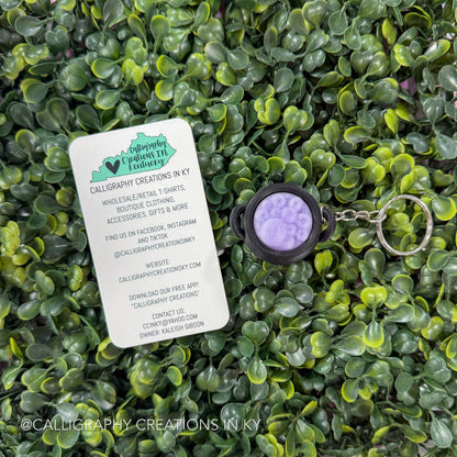 A calligraphy-themed keychain with a round purple and lavender design in a black bezel, shown next to a white brand card on a background of green artificial leaves.