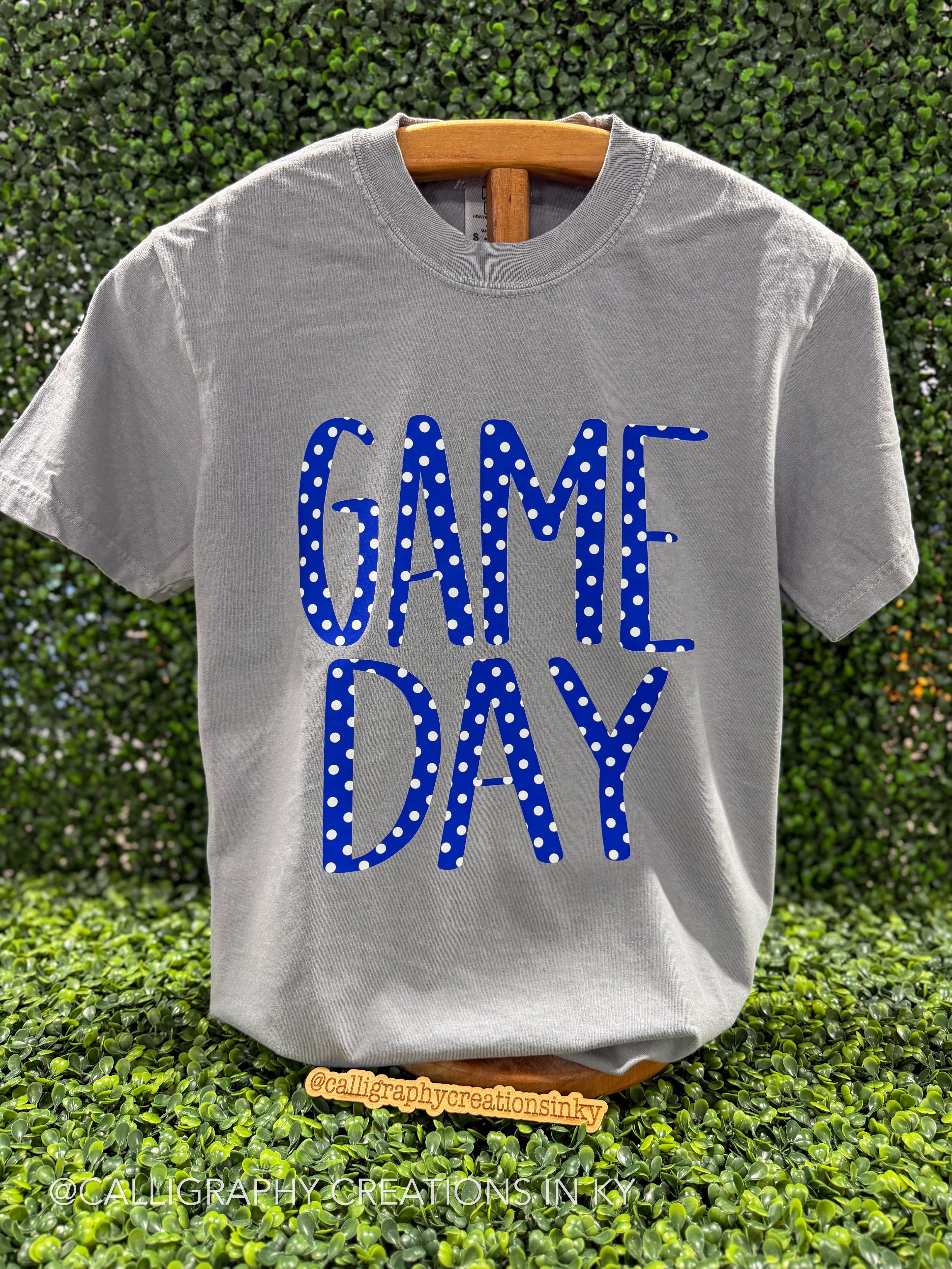 A light gray crewneck t-shirt on a wooden hanger, featuring the words 'GAME DAY' in blue block letters filled with white polka dots, set against a green hedge background.