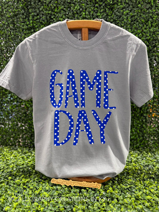 A light gray crewneck t-shirt on a wooden hanger, featuring the words 'GAME DAY' in blue block letters filled with white polka dots, set against a green hedge background.