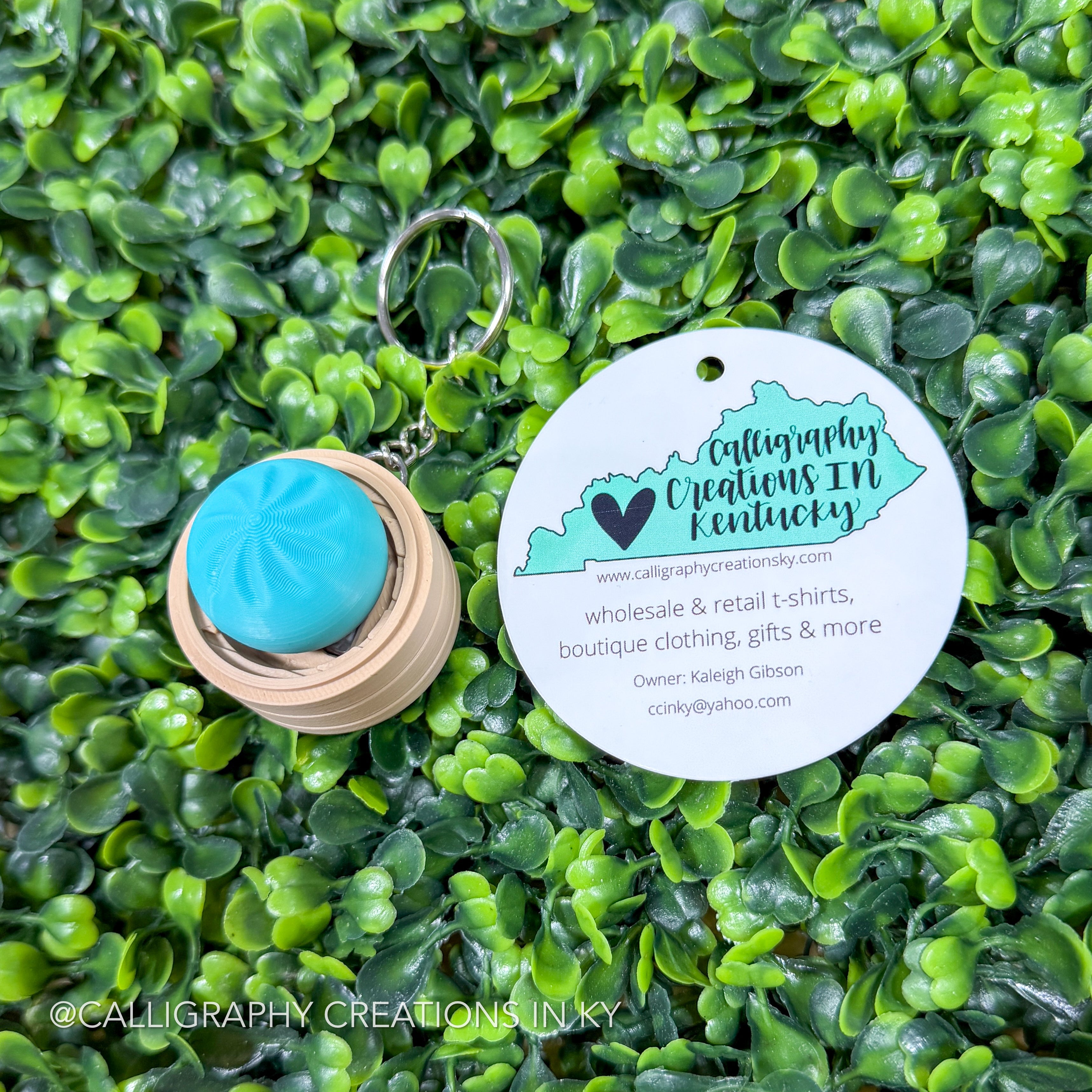 Keychain with teal gradient dumpling fidget clicker and 'Calligraphy Creations in Kentucky' tag on green foliage