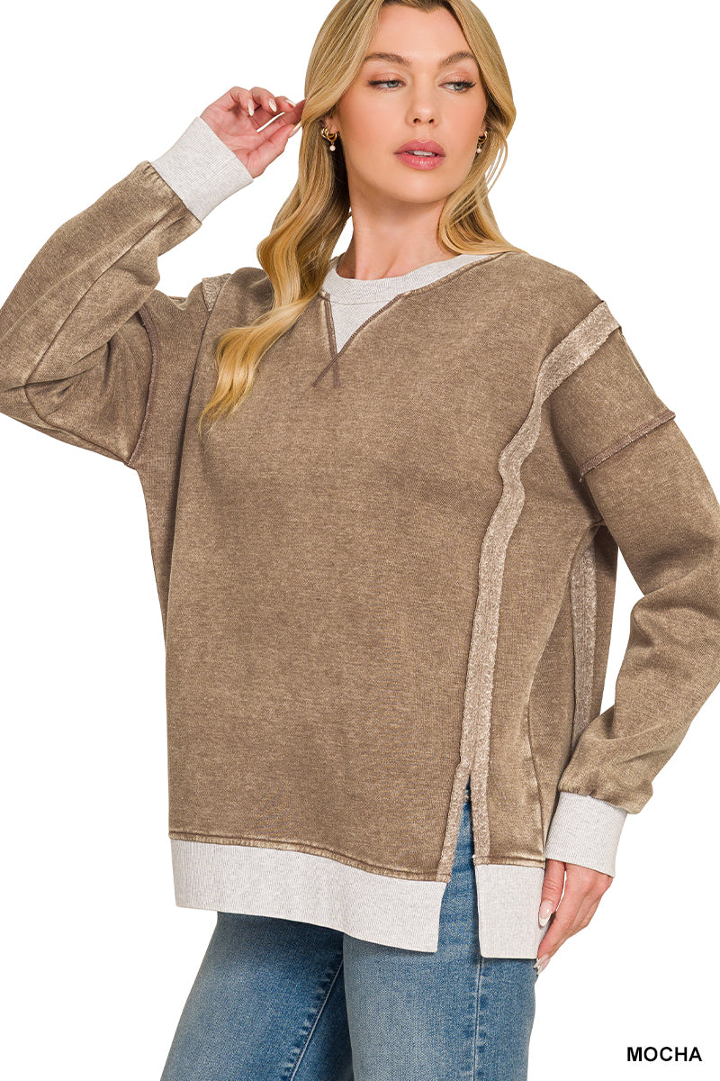 A woman modeling a long-sleeve V-neck sweater in a heathered mocha brown color, featuring contrasting light gray ribbed trim at the collar, cuffs, hem, and side panels, paired with blue jeans against a white background.