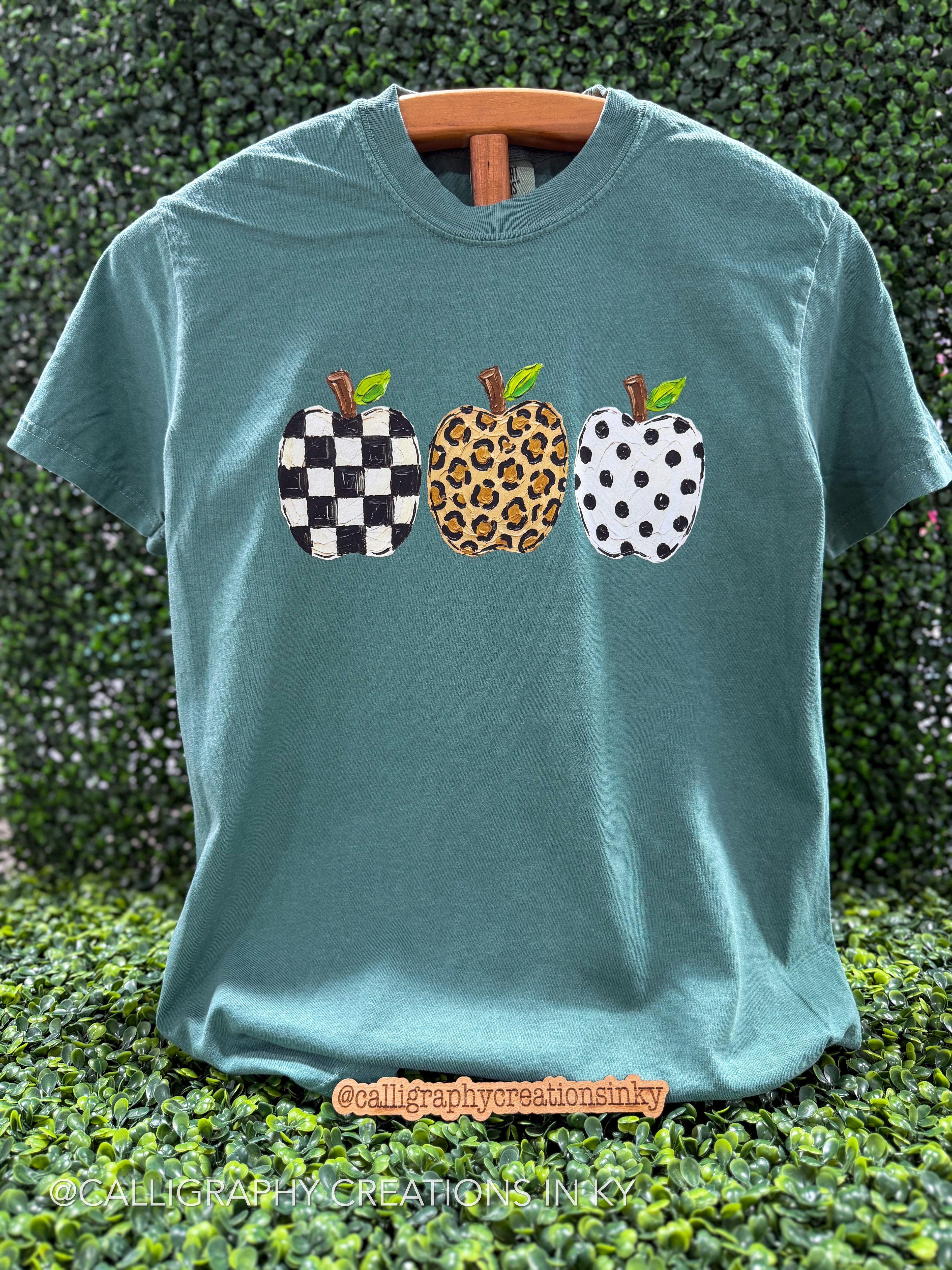 A teal short-sleeve t-shirt with three decorative apple graphics on the chest, displayed on a wooden hanger against a green hedge background. The apples feature black-and-white checkered, leopard print, and white-and-black polka dot patterns.