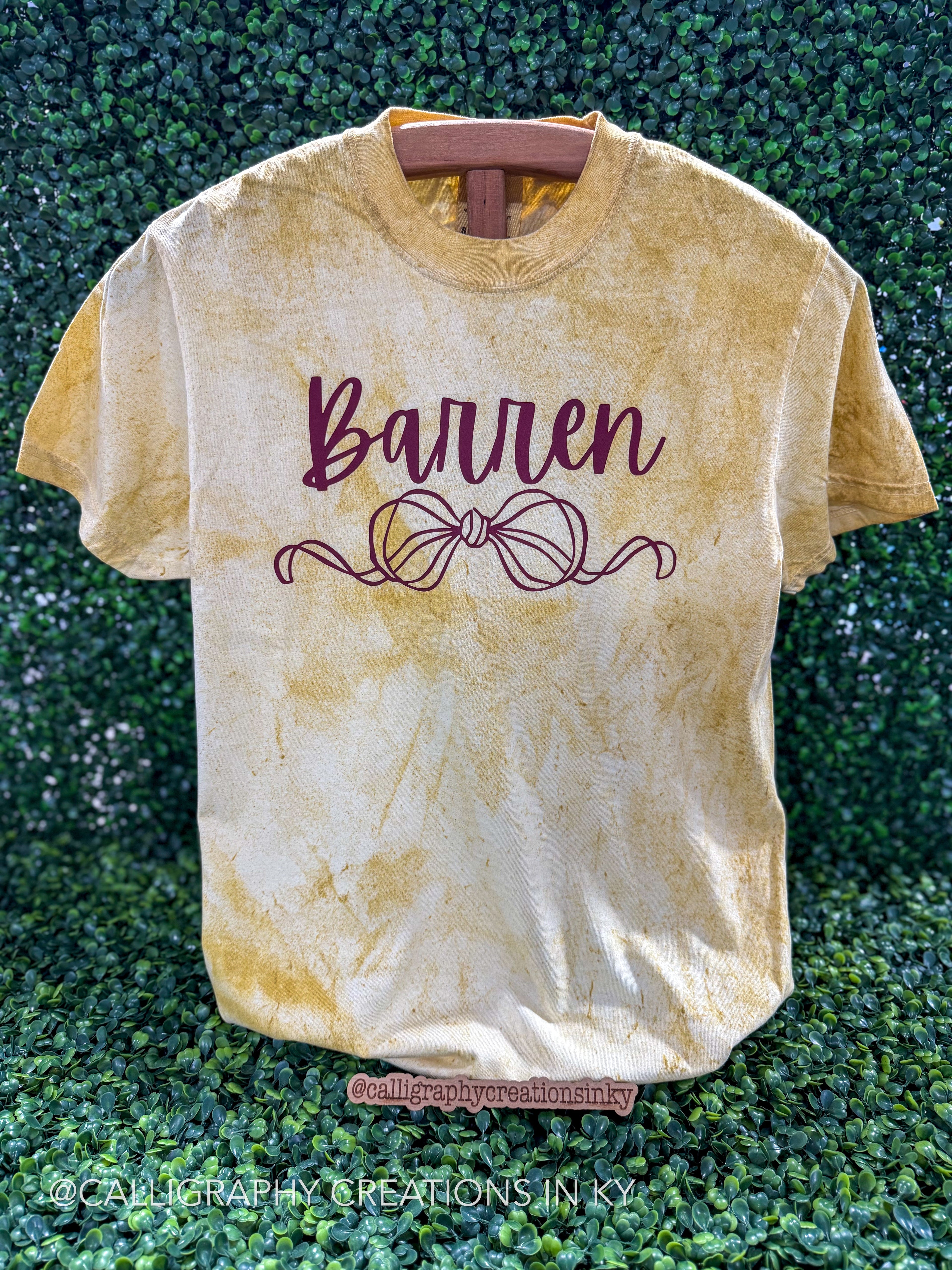 A short-sleeve crewneck t-shirt with a yellow and cream mineral-wash finish, featuring the name 'Barren' and a bow design in dark burgundy on the front, displayed on a wooden hanger against a green hedge background.