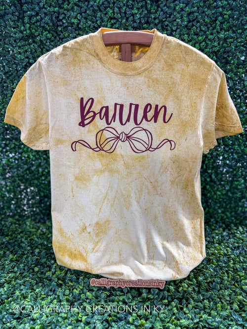 A short-sleeve crewneck t-shirt with a yellow and cream mineral-wash finish, featuring the name 'Barren' and a bow design in dark burgundy on the front, displayed on a wooden hanger against a green hedge background.