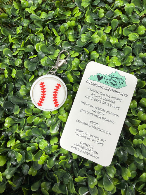 A baseball-themed keychain with a white circular charm featuring red stitching details and a silver clasp, displayed on a bed of green artificial foliage next to its white paper tag.