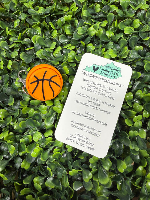 A small orange basketball-shaped keychain with black basketball lines lies on a white printed card, which is placed on dense green foliage. The card shows the brand name Caliber Keychain Designs in Ky.