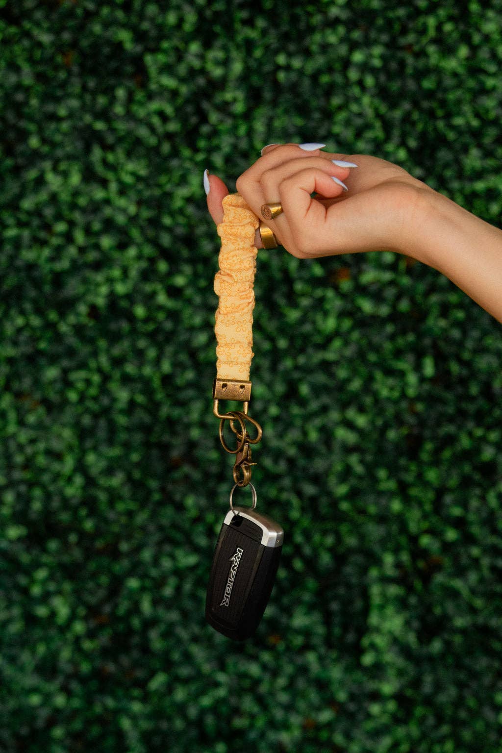 Hand holding a keychain with a yellow scrunchie keychain against a green hedge background