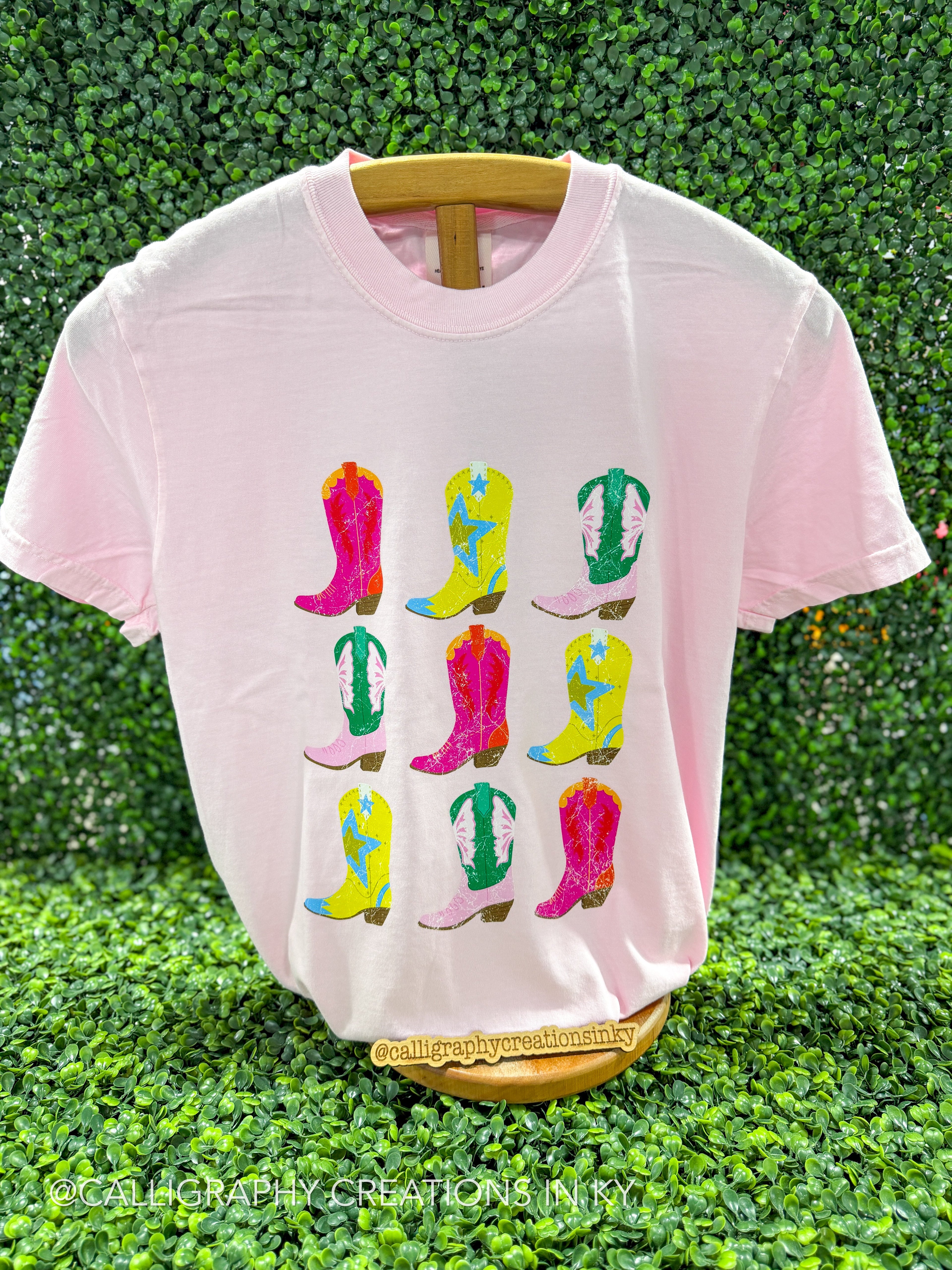 A light pink short-sleeve t-shirt with a graphic print of nine colorful cowboy boots arranged in a 3x3 grid, displayed on a wooden hanger against a green leaf wall background.