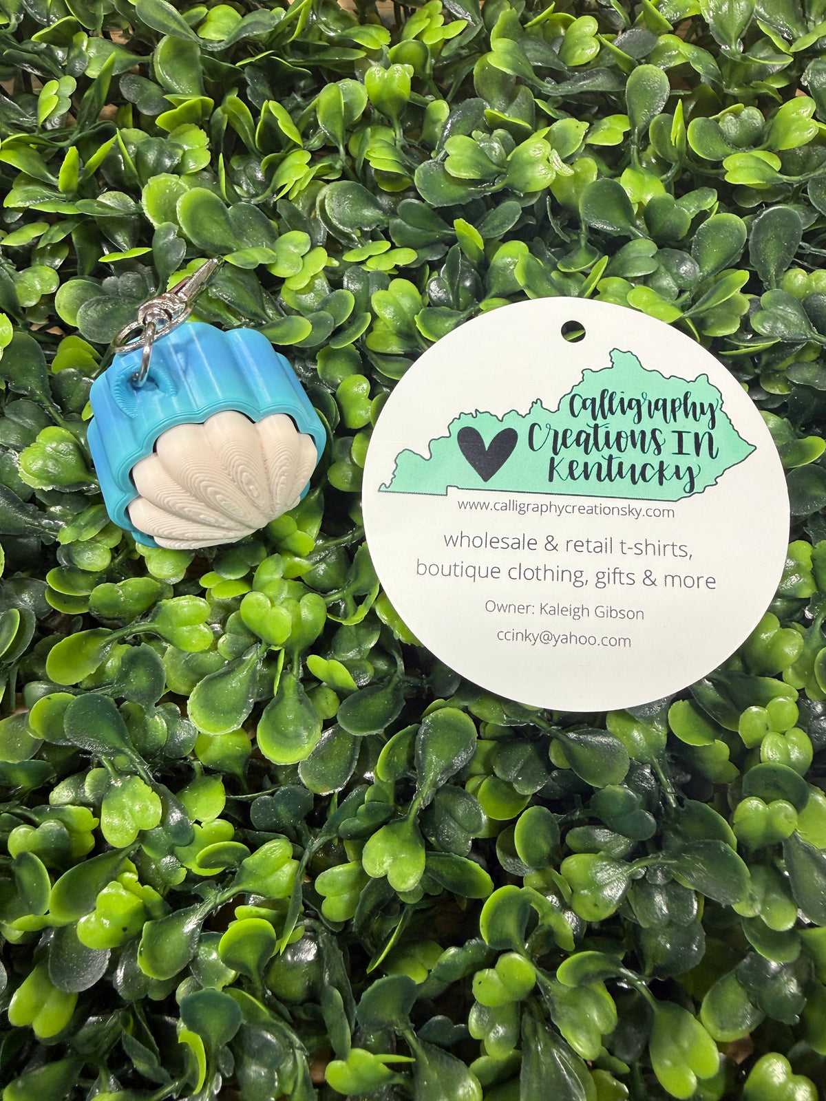 Blue and white shell-shaped keychain on green leafy background with a visible brand label.