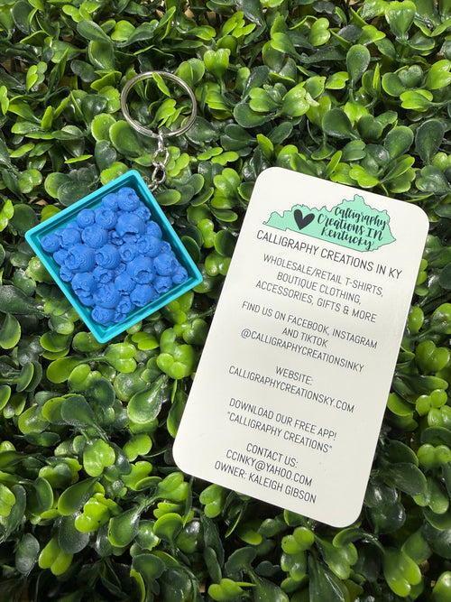 A small square turquoise container filled with bright blue flower-shaped calligraphy charms, placed on green foliage next to a white printed card with branding and contact details.