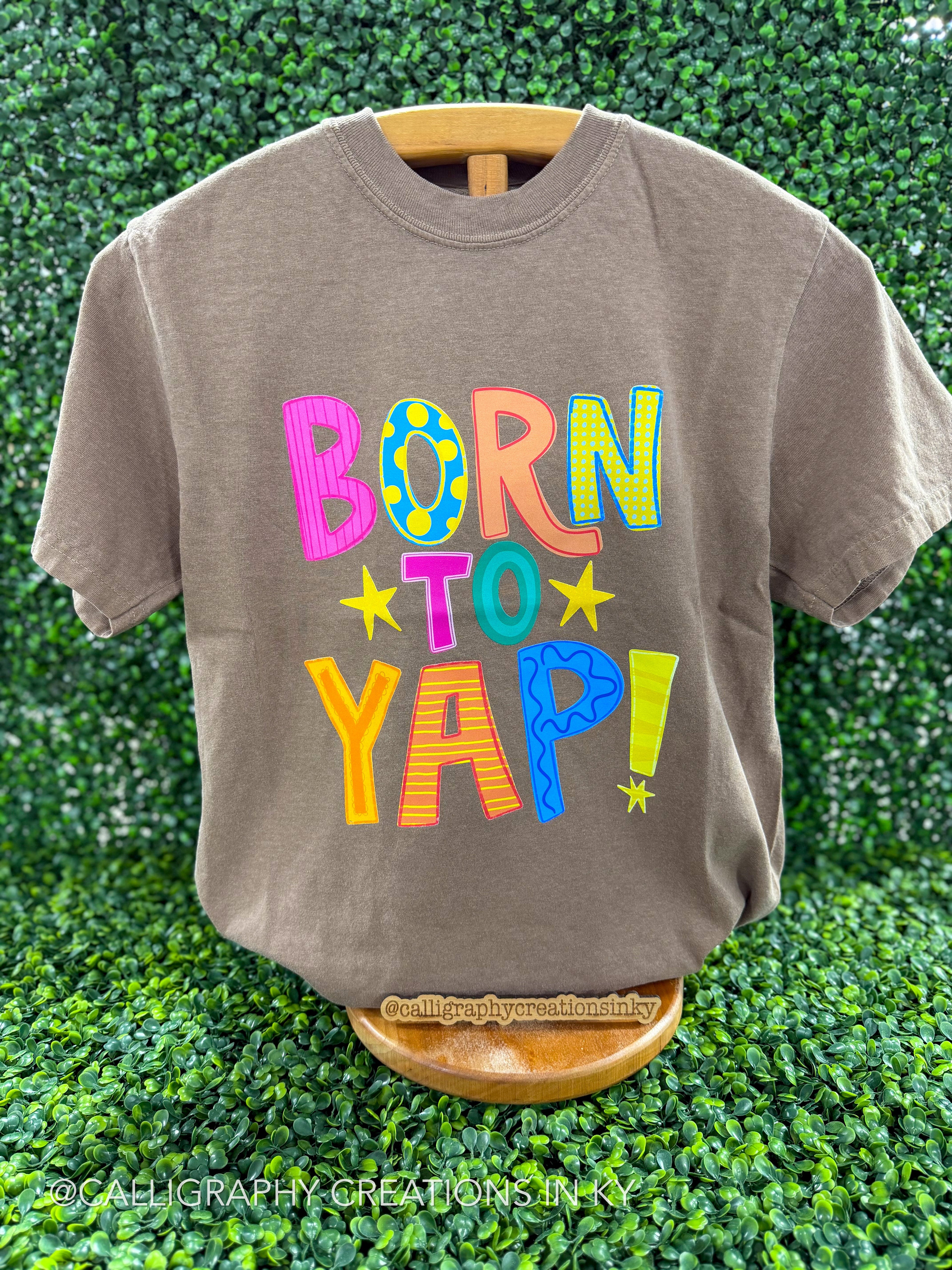 Brown shirt with colorful patterned letters that spell our ‘Born to yap’ on a wooden stand