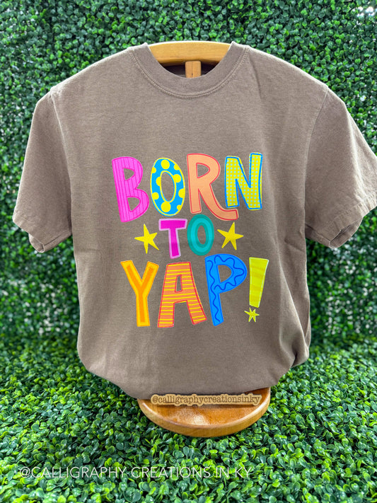 Brown shirt with colorful patterned letters that spell our ‘Born to yap’ on a wooden stand