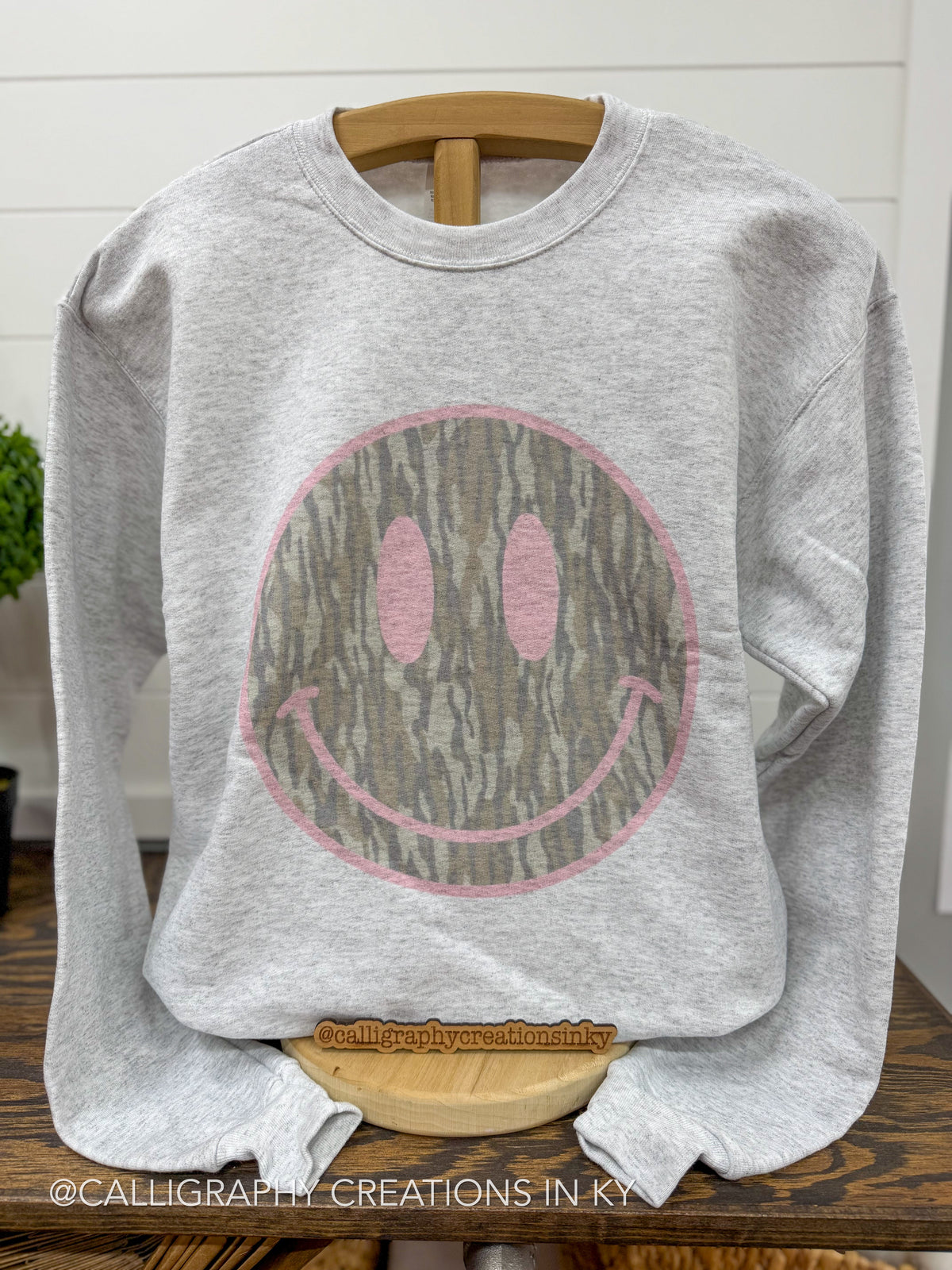 A light heather gray crewneck sweatshirt displayed on a wooden hanger. The sweatshirt features a large smiley face graphic on the front, where the smiley face is filled with a brown and gray tree bark pattern and outlined in pink.