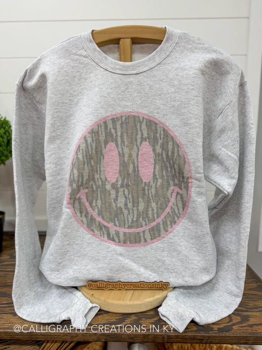 A light heather gray crewneck sweatshirt displayed on a wooden hanger. The sweatshirt features a large smiley face graphic on the front, where the smiley face is filled with a brown and gray tree bark pattern and outlined in pink.