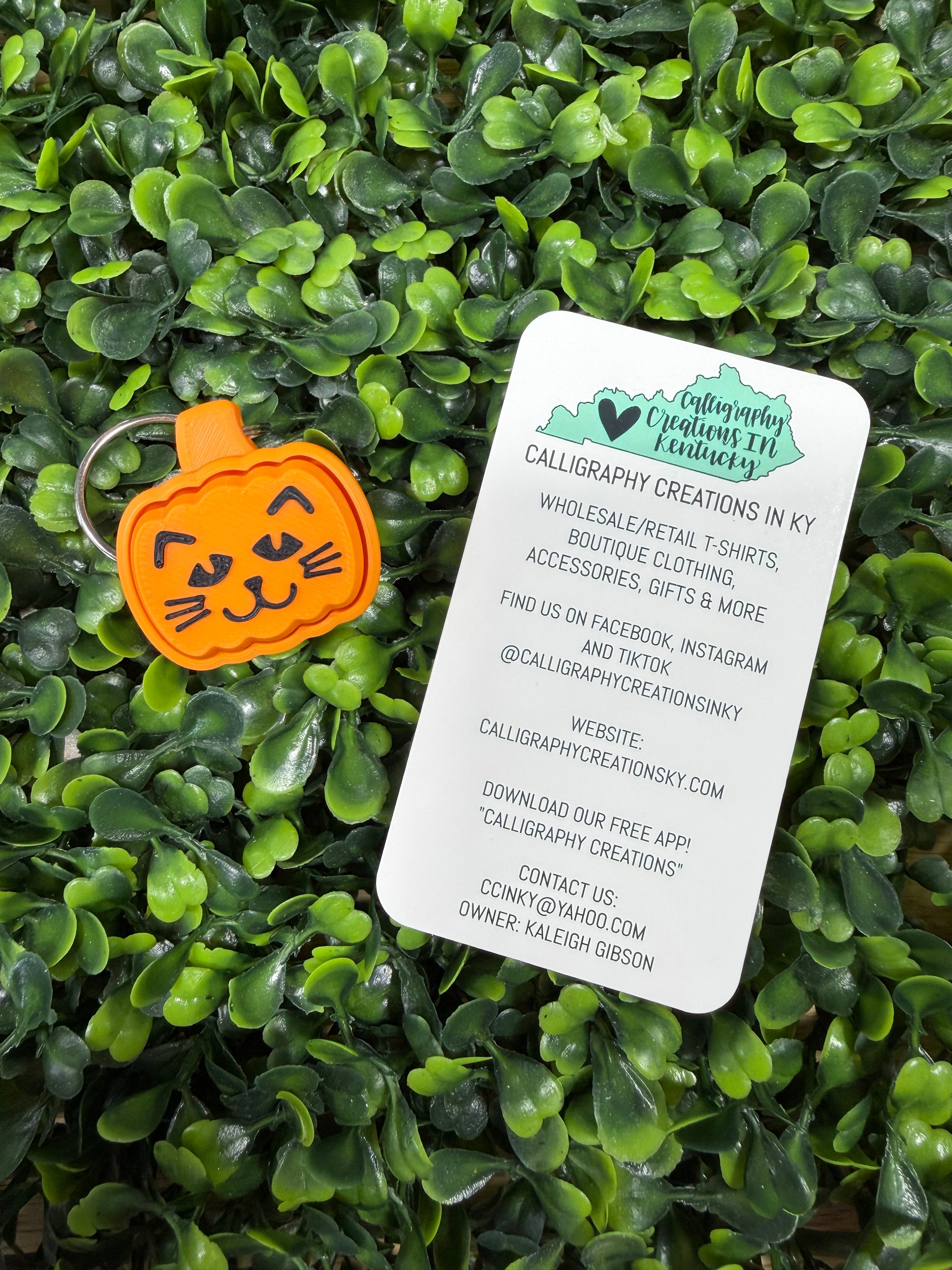 An orange pumpkin-shaped calligraphy keychain with a cat face sits on a white branded card, placed on a background of dense green artificial foliage.