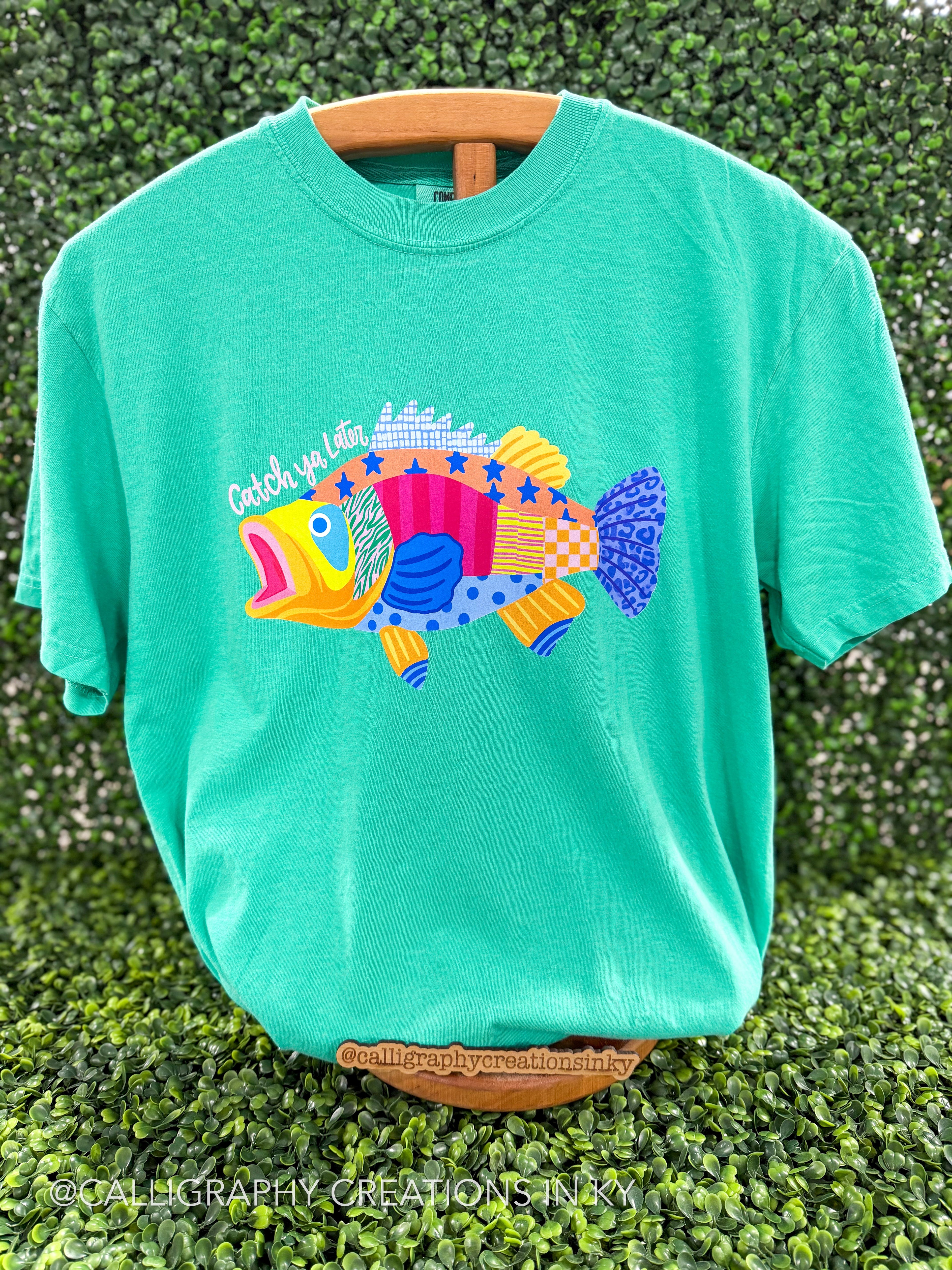 Green shirt with patterned bass that says ‘Catch ya Later’ on a wooden stand