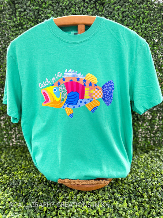 Green shirt with patterned bass that says ‘Catch ya Later’ on a wooden stand