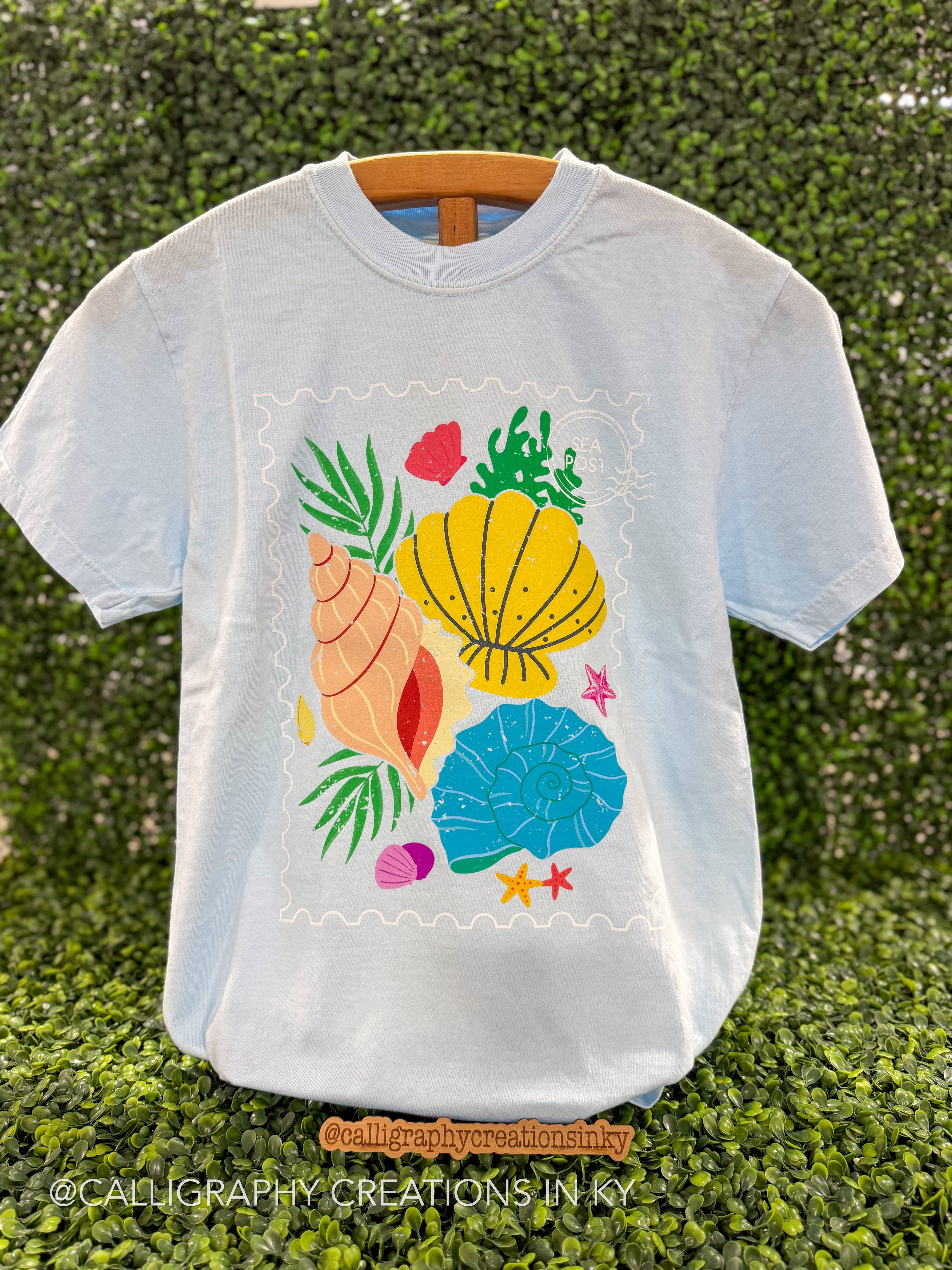 Light blue t shirt with colorful shells printed on a wooden stand 