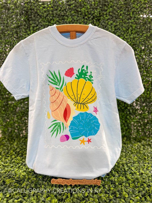 Light blue t shirt with colorful shells printed on a wooden stand 