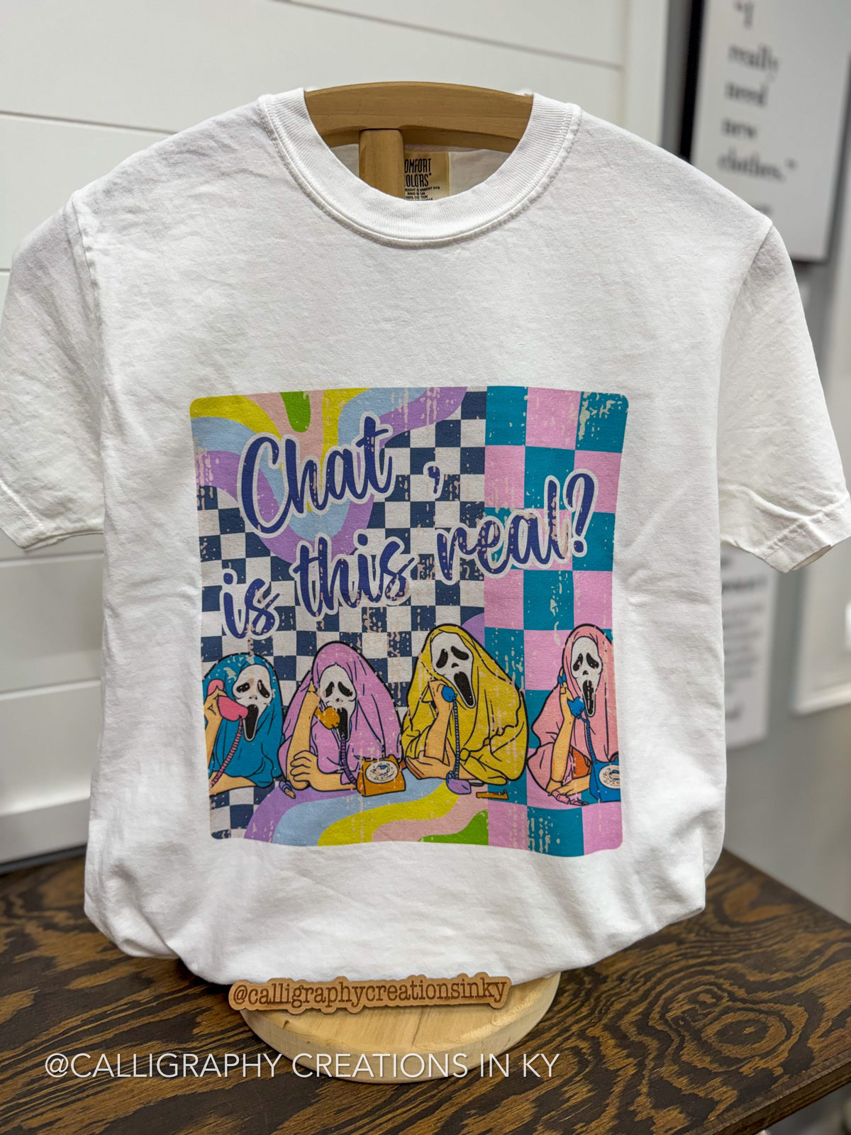 A white short-sleeve graphic T-shirt hanging on a wooden hanger against a white paneled wall. The shirt features a colorful front print with the text 'Chat is this real?' above four cartoon ghosts sitting on a checkered pastel background.