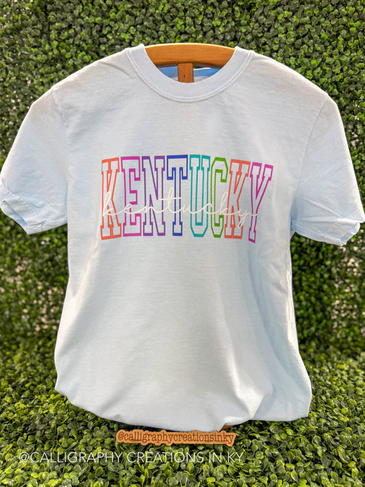 Blue shirt with rainbow Kentucky letters with script font over top on a wooden stand 