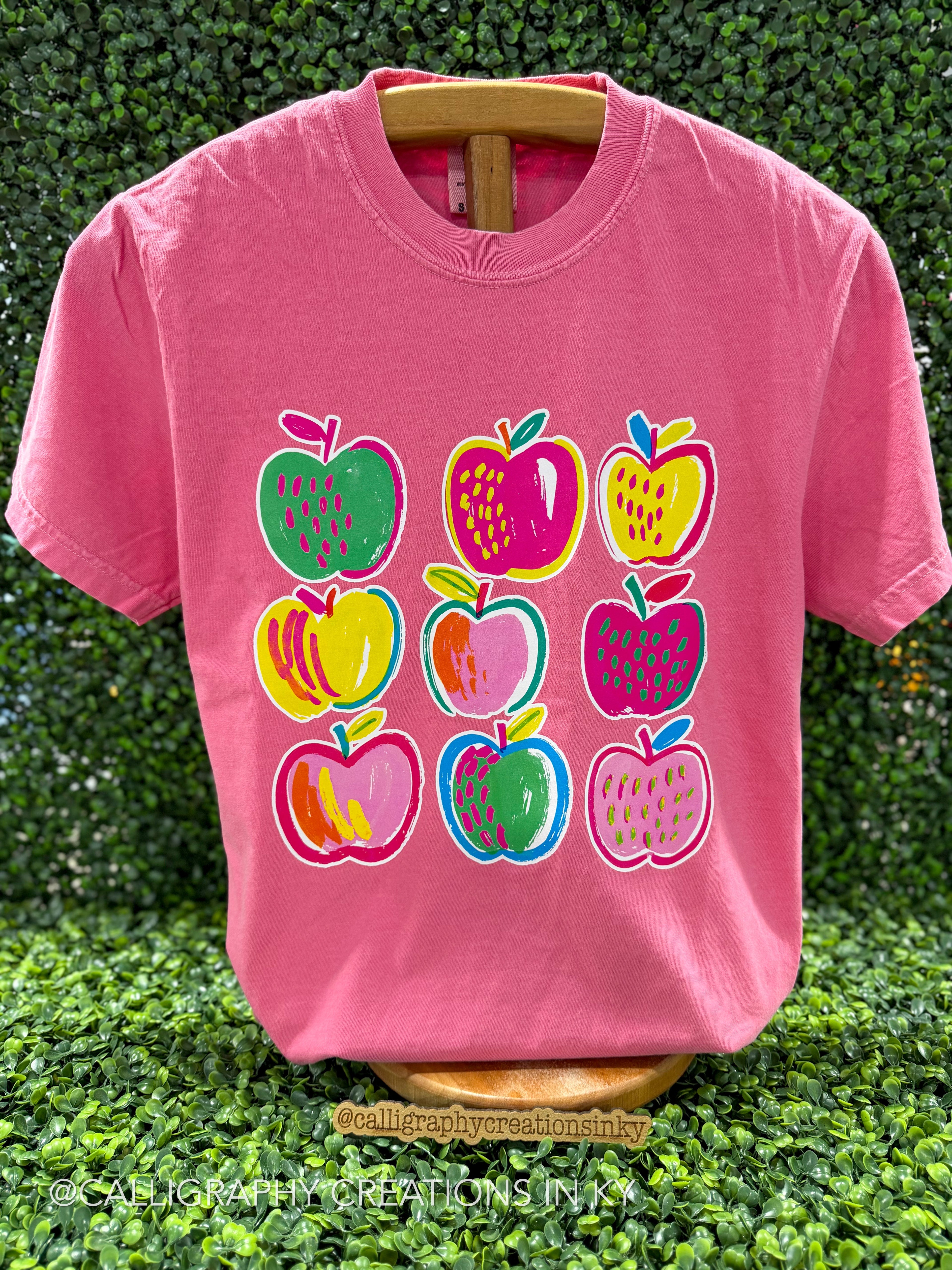 A pink short-sleeve t-shirt displayed on a wooden hanger against a green hedge background. The front of the shirt features a graphic print of nine colorful apples in various patterns and colors, including green, pink, yellow, and blue.