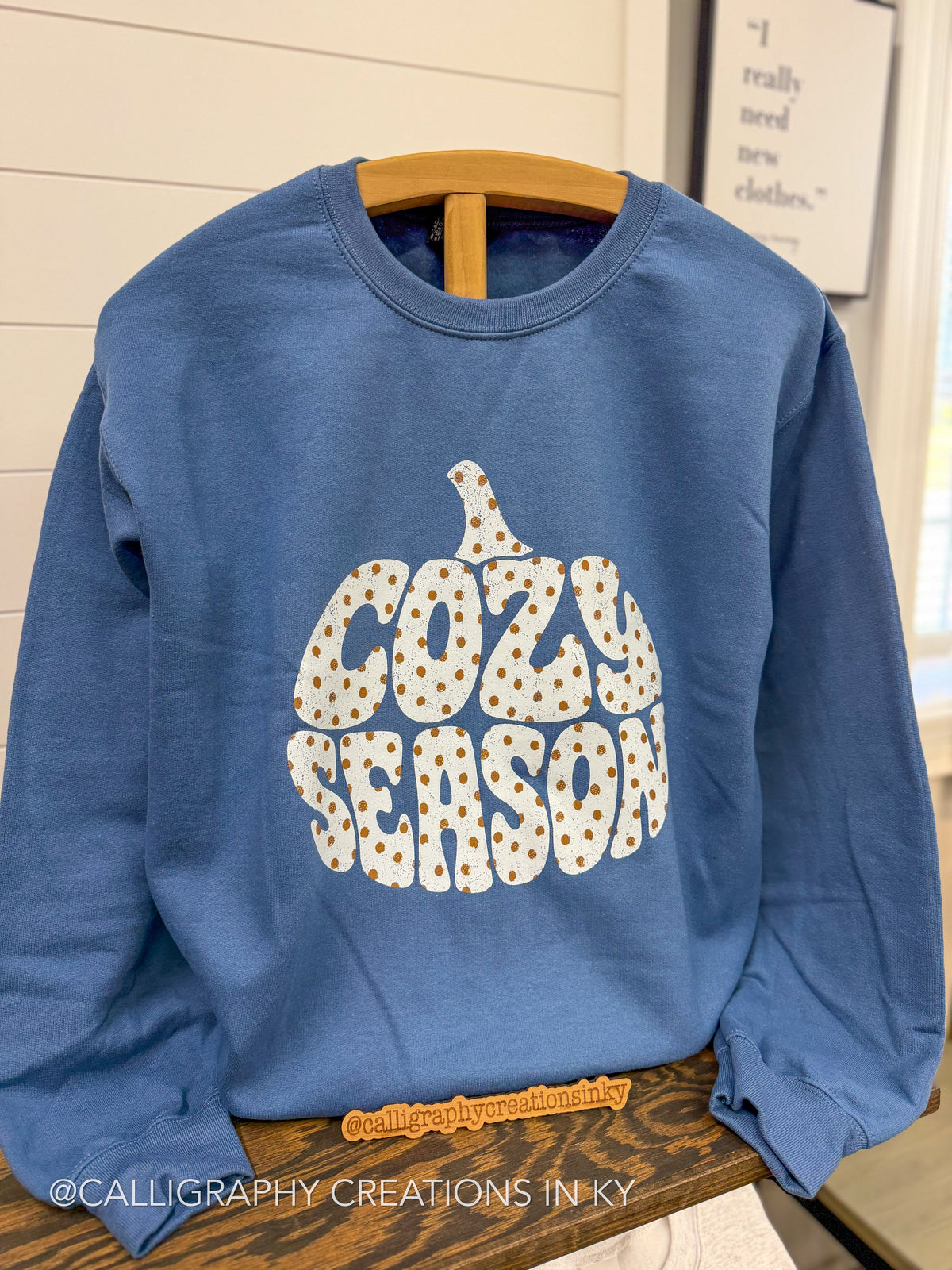 A blue crewneck sweatshirt with a pumpkin-shaped graphic reading 'COZY SEASON' in white letters filled with small orange polka dots, hanging on a wooden hanger.