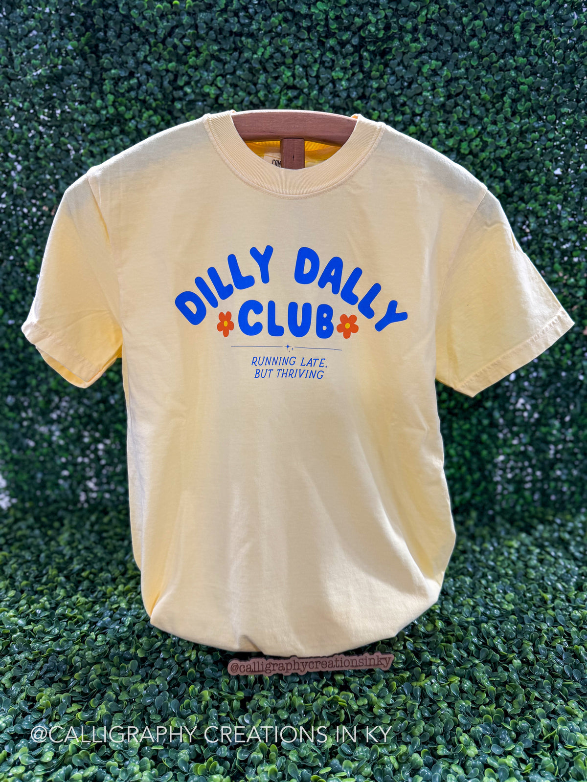 A pale yellow t-shirt with blue and orange graphic text reading 'DILLY DAILY CLUB' and 'RUNNING LATE, BUT THRIVING' displayed against a green hedge background.