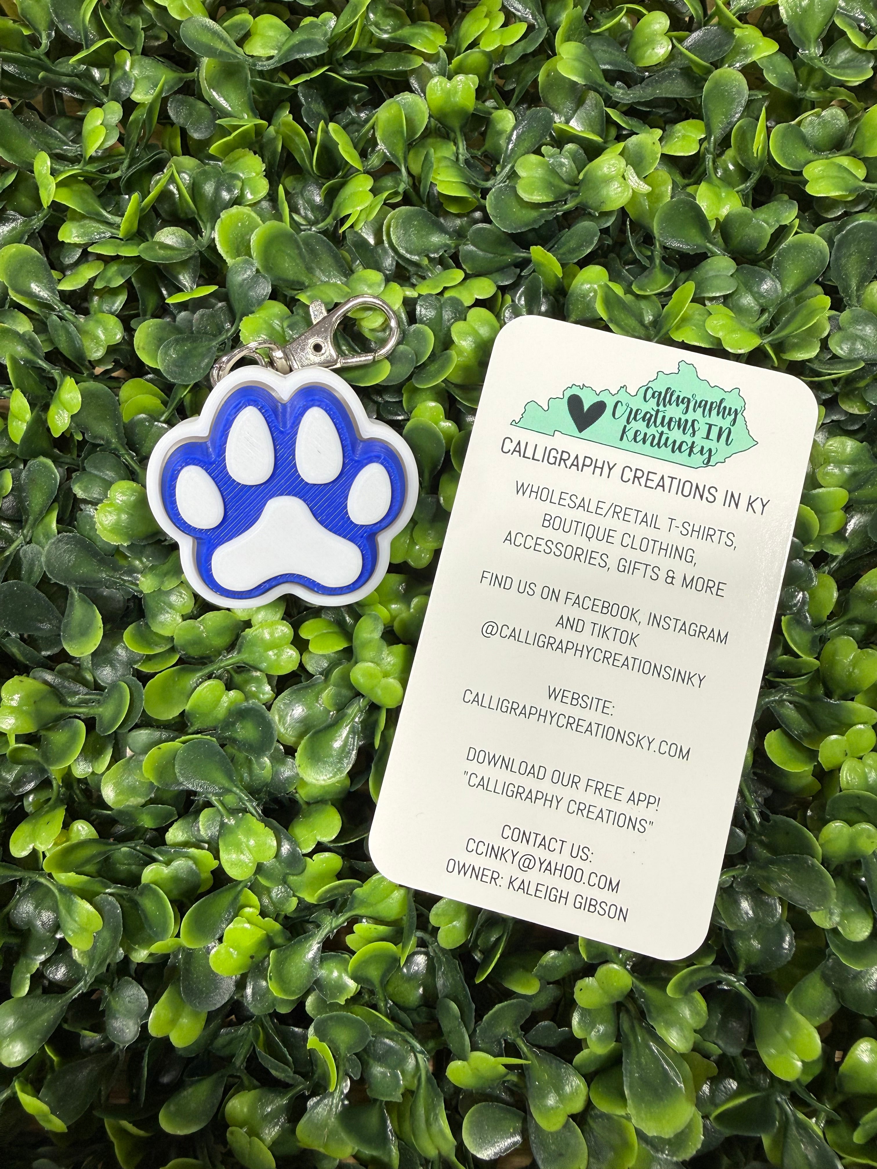 A blue and white paw print pet tag with a silver keyring, displayed on green artificial foliage next to a white paper tag with branding details.
