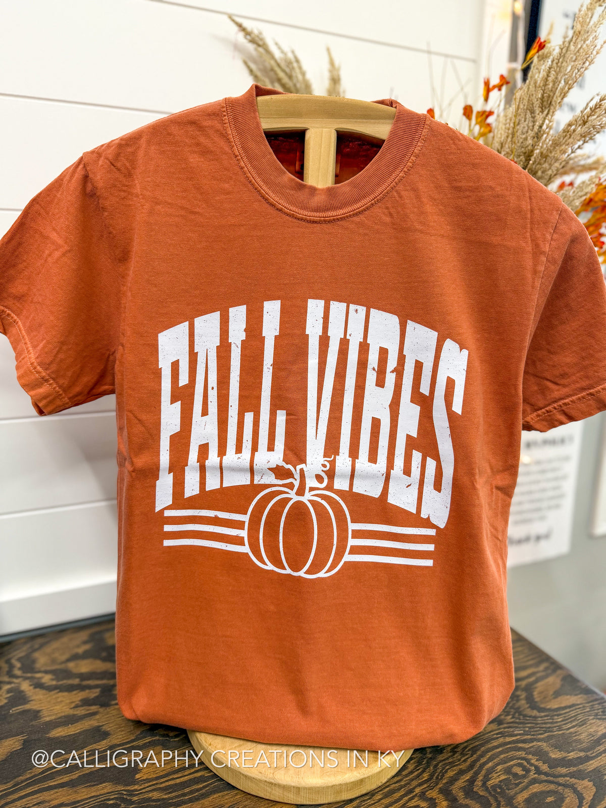 An orange short-sleeve t-shirt displayed on a wooden hanger, featuring a white graphic print that reads 'FALL VIBES' above a pumpkin illustration. The shirt is photographed in a styled indoor setting with fall decorations in the background.