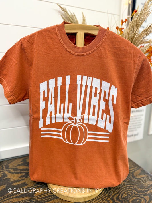 An orange short-sleeve t-shirt displayed on a wooden hanger, featuring a white graphic print that reads 'FALL VIBES' above a pumpkin illustration. The shirt is photographed in a styled indoor setting with fall decorations in the background.