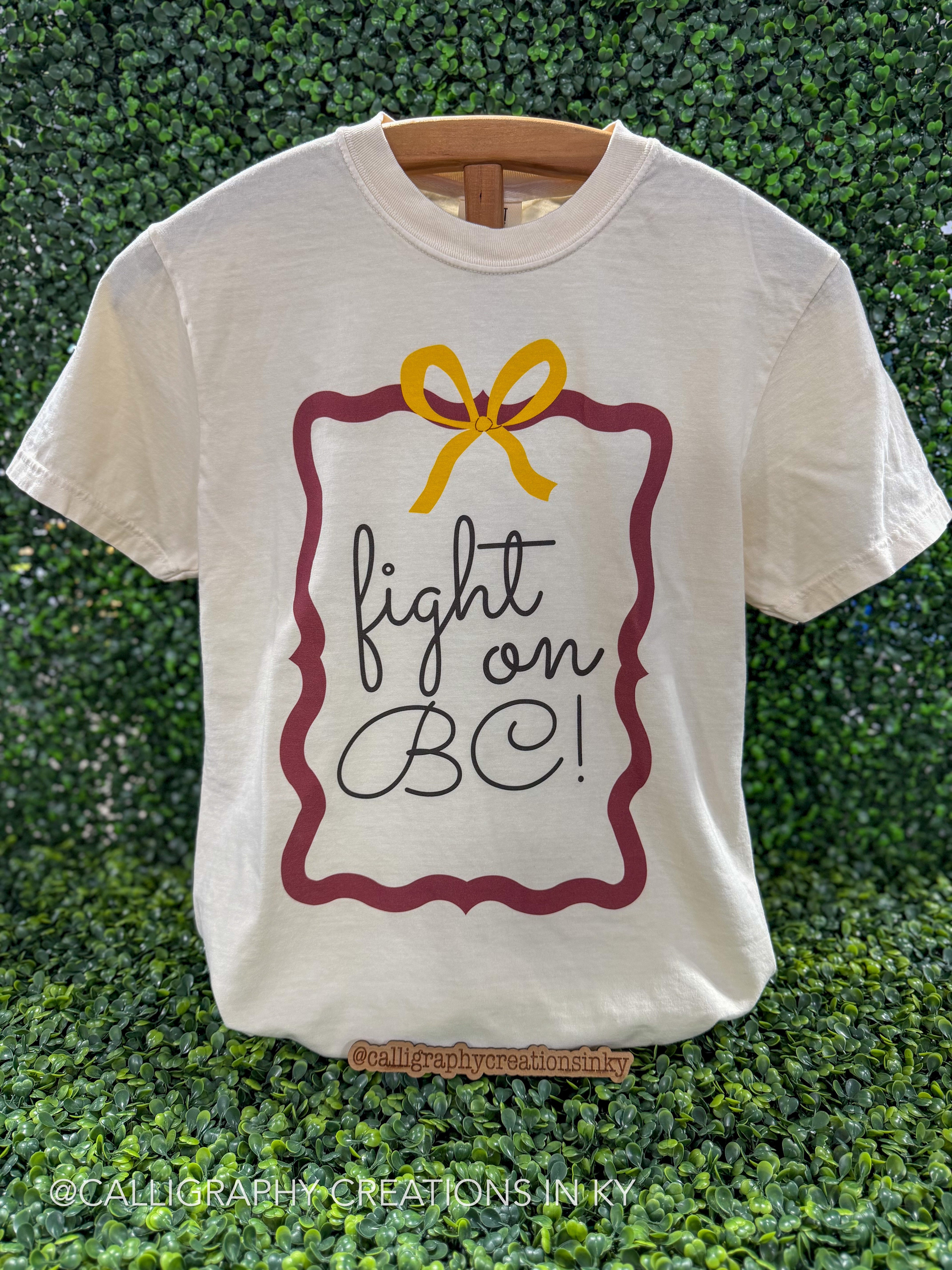 An off-white short-sleeve t-shirt on a wooden hanger, featuring a graphic print with a maroon border, a yellow bow, and the text 'fight on BC!' in black script, set against a green leaf wall background.