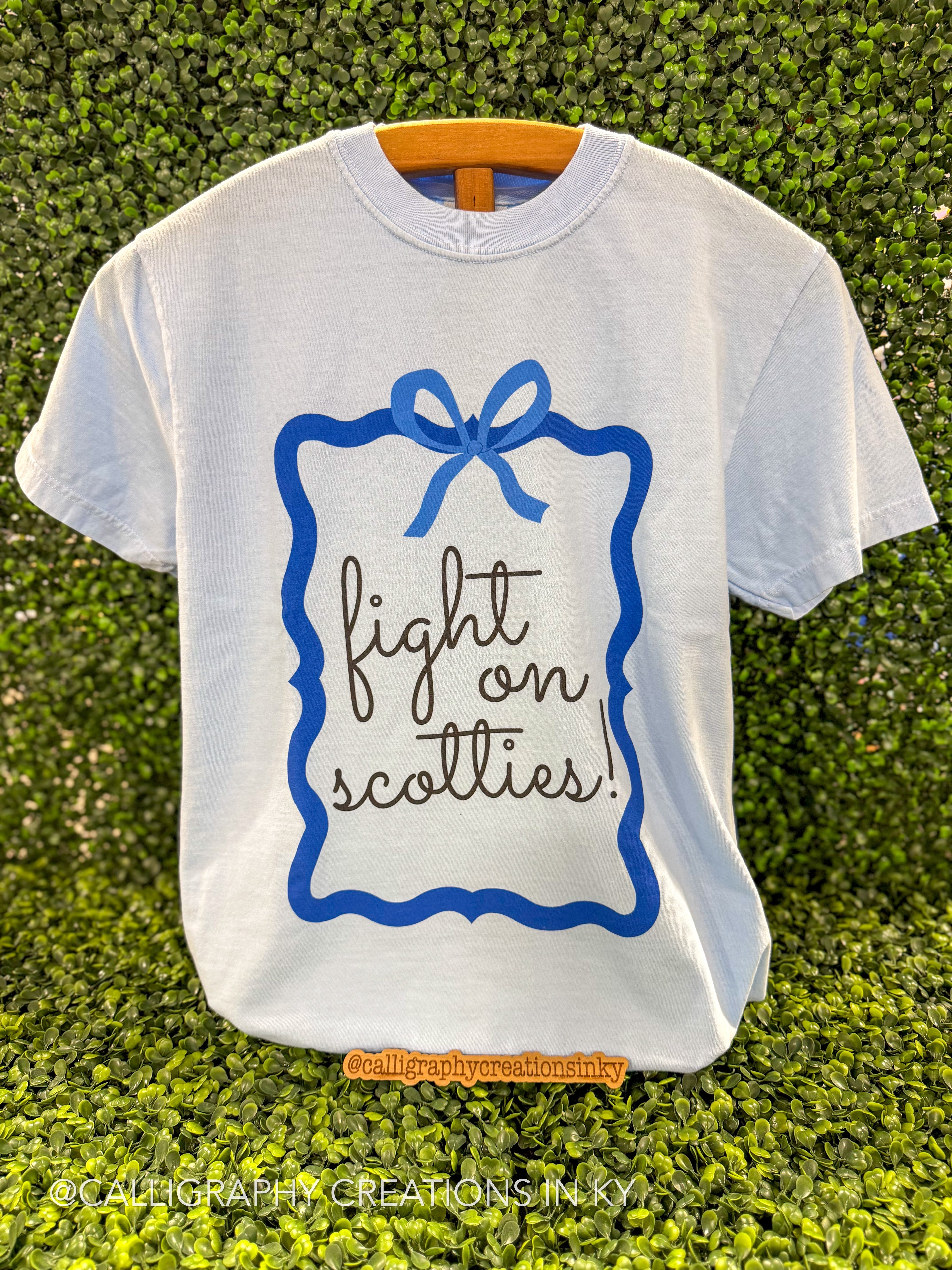 A white short-sleeve t-shirt with a blue decorative border and bow graphic, featuring the text 'fight on scotties!' in black cursive and print. The shirt is displayed on a wooden hanger against a green leafy wall background.