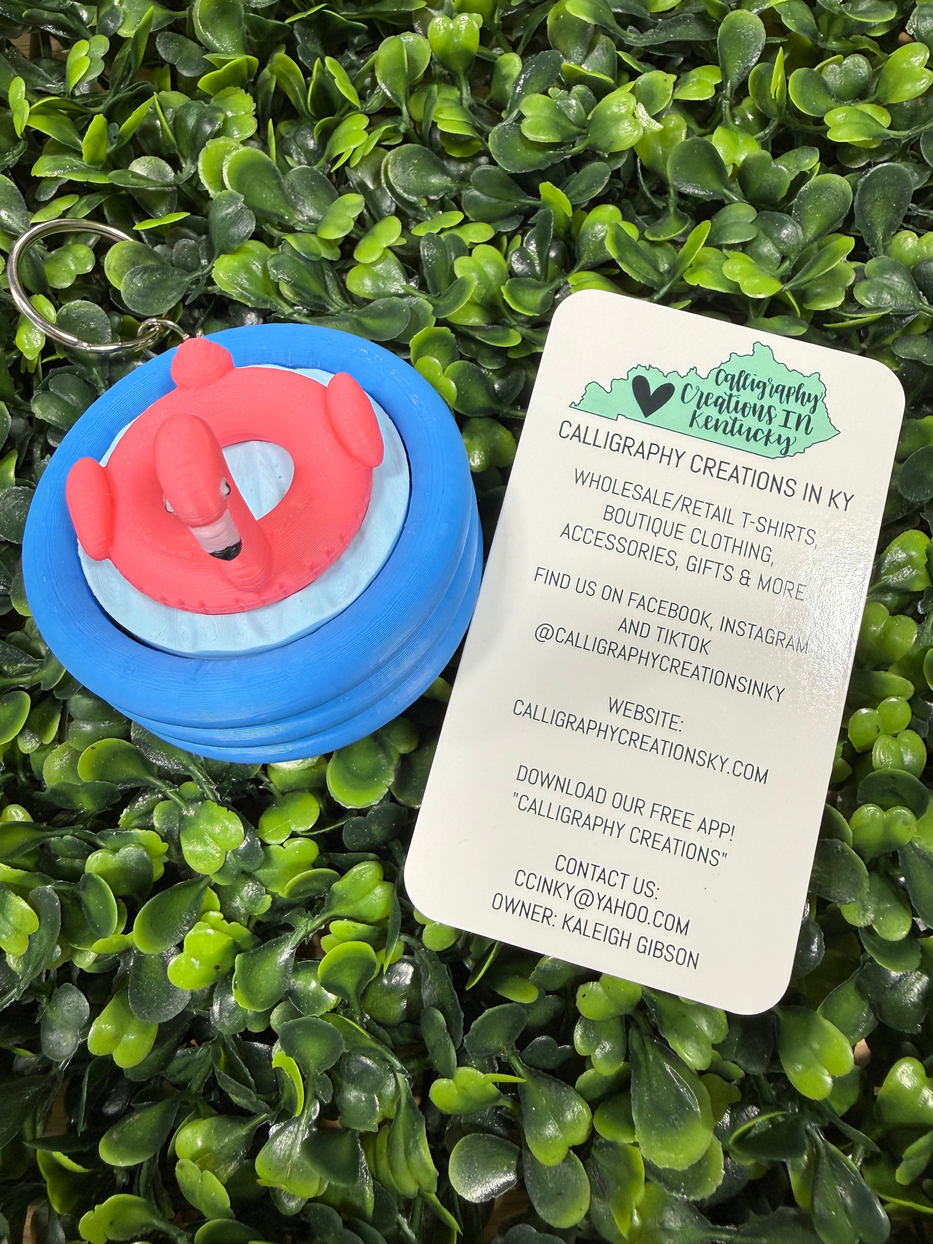 A small blue and pink flamingo pool floatie keychain charm lies on a white paper tag with brand information, placed on a bed of green artificial leaves.