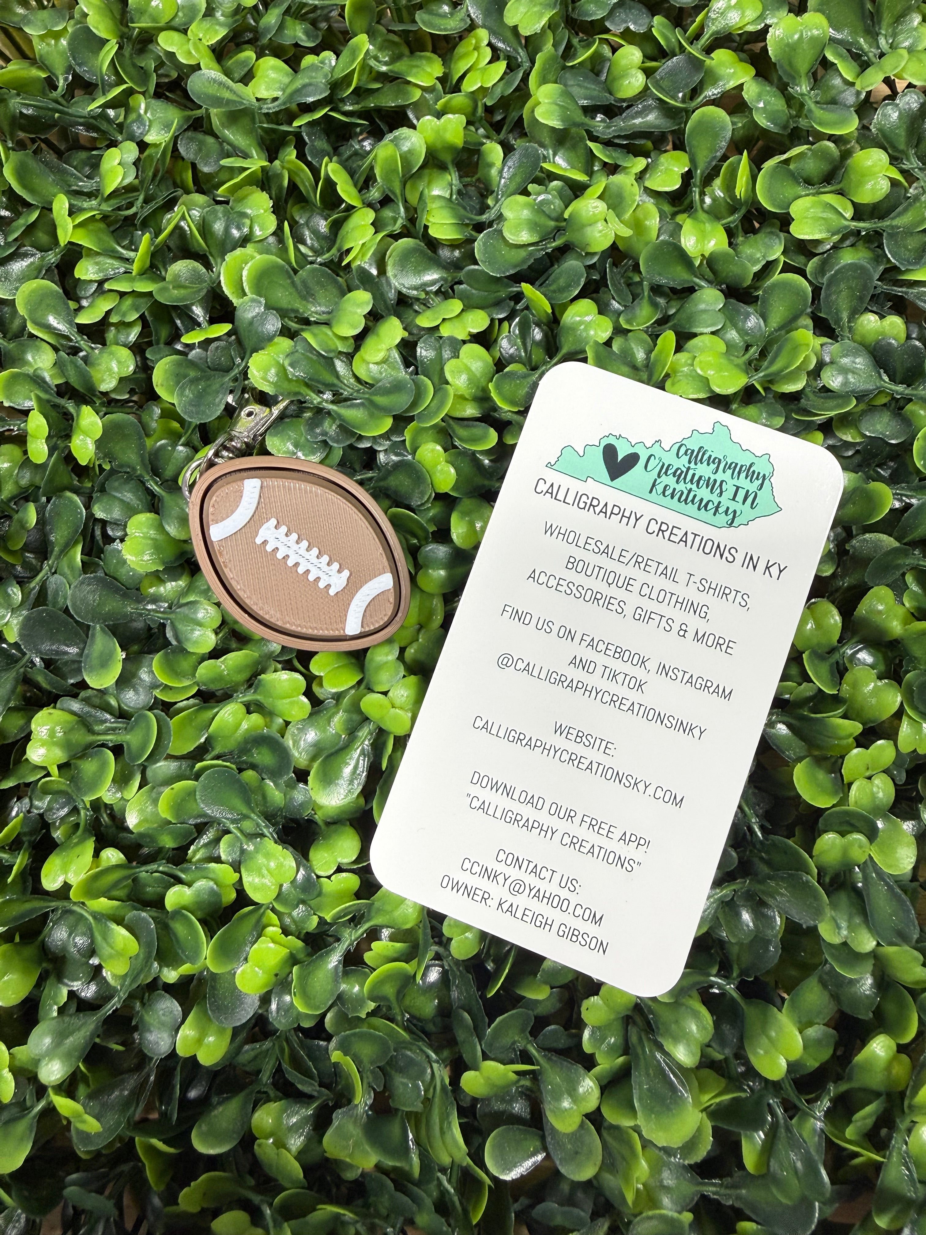 A football-shaped keychain with a brown leather-look body and white laces, displayed on a bed of green artificial leaves next to a white product tag.