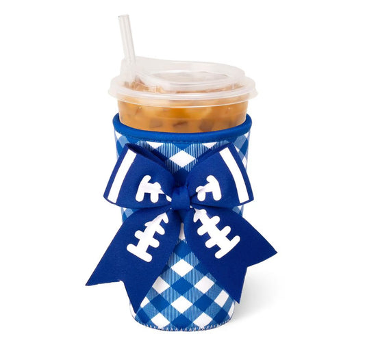A blue and white gingham football-themed cup sleeve with a large blue bow printed with white football laces, holding an iced coffee in a clear plastic cup with a straw, on a white background.