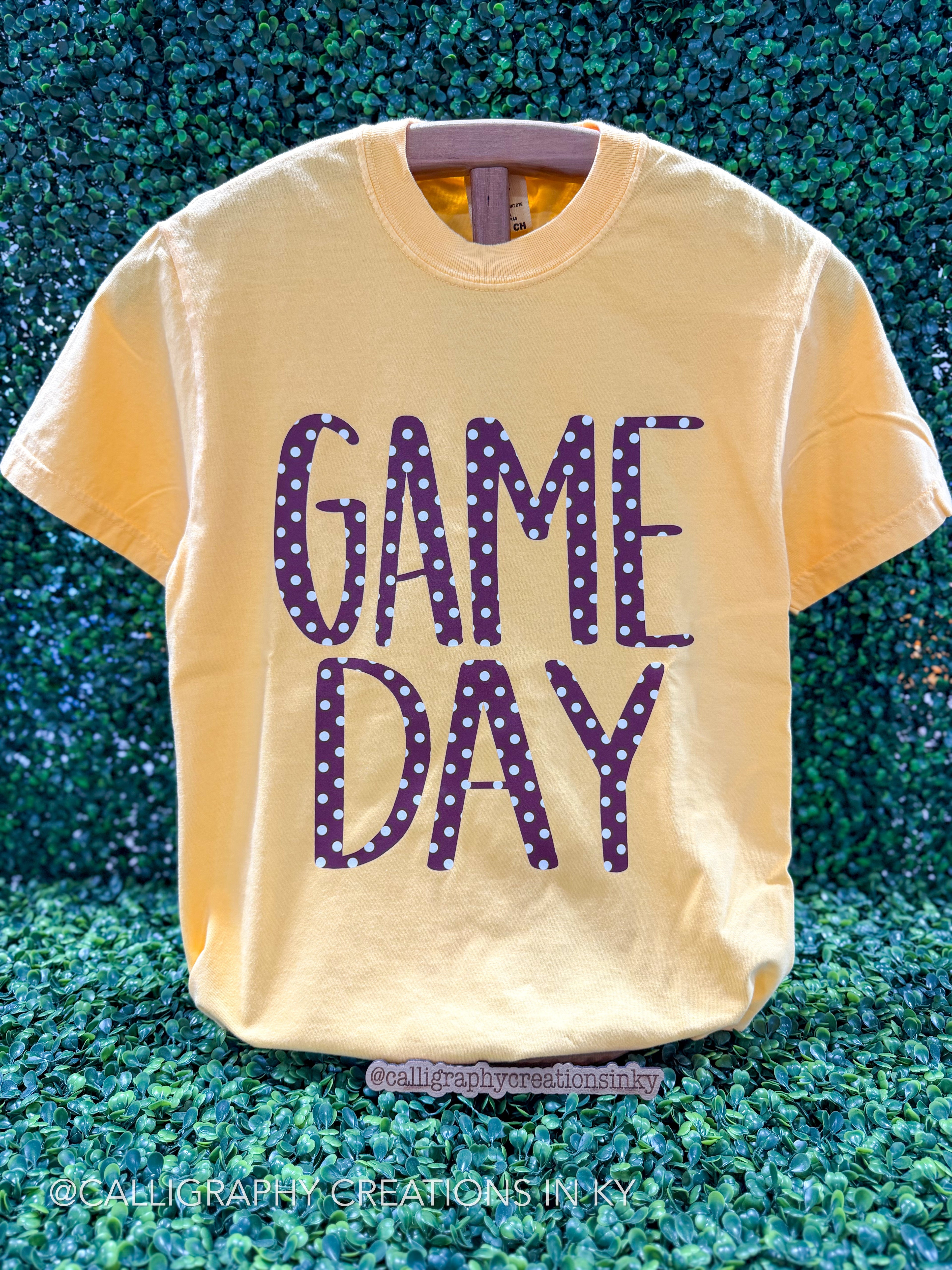 A pale yellow graphic t-shirt featuring the words 'GAME DAY' in a purple and white polka-dot patterned font, displayed against a green hedge background.