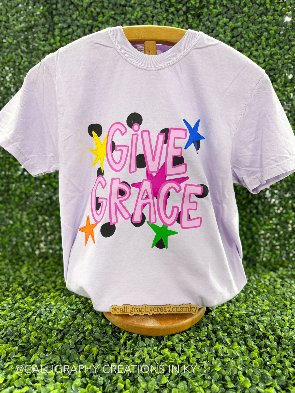 Light purple shirt that says ‘Give Grace’ with stars on a wooden stand