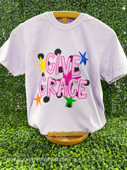 Light purple shirt that says ‘Give Grace’ with stars on a wooden stand