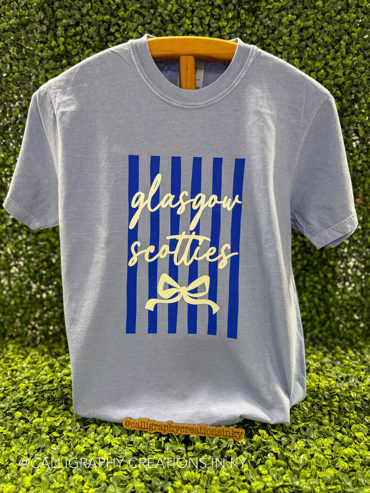 A light gray crewneck t-shirt hanging on a wooden hanger against a green hedge background. The shirt features a graphic print with vertical blue stripes, the words 'glasgow scotties' in cream cursive, and a cream bow illustration.