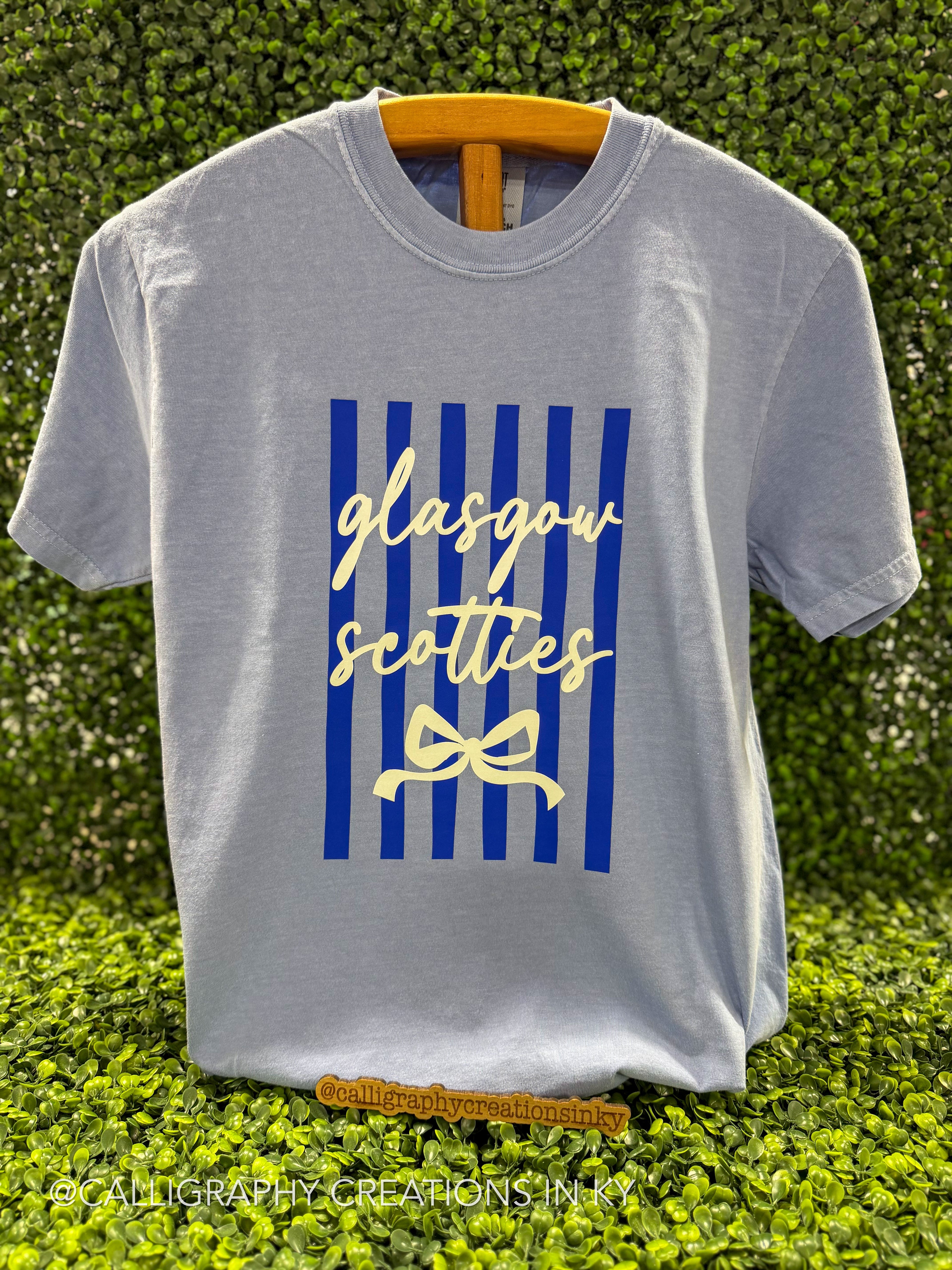 A light gray crewneck t-shirt hanging on a wooden hanger against a green hedge background. The shirt features a graphic print with vertical blue stripes, the words 'glasgow scotties' in cream cursive, and a cream bow illustration.