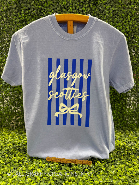 A light gray crewneck t-shirt hanging on a wooden hanger against a green hedge background. The shirt features a graphic print with vertical blue stripes, the words 'glasgow scotties' in cream cursive, and a cream bow illustration.